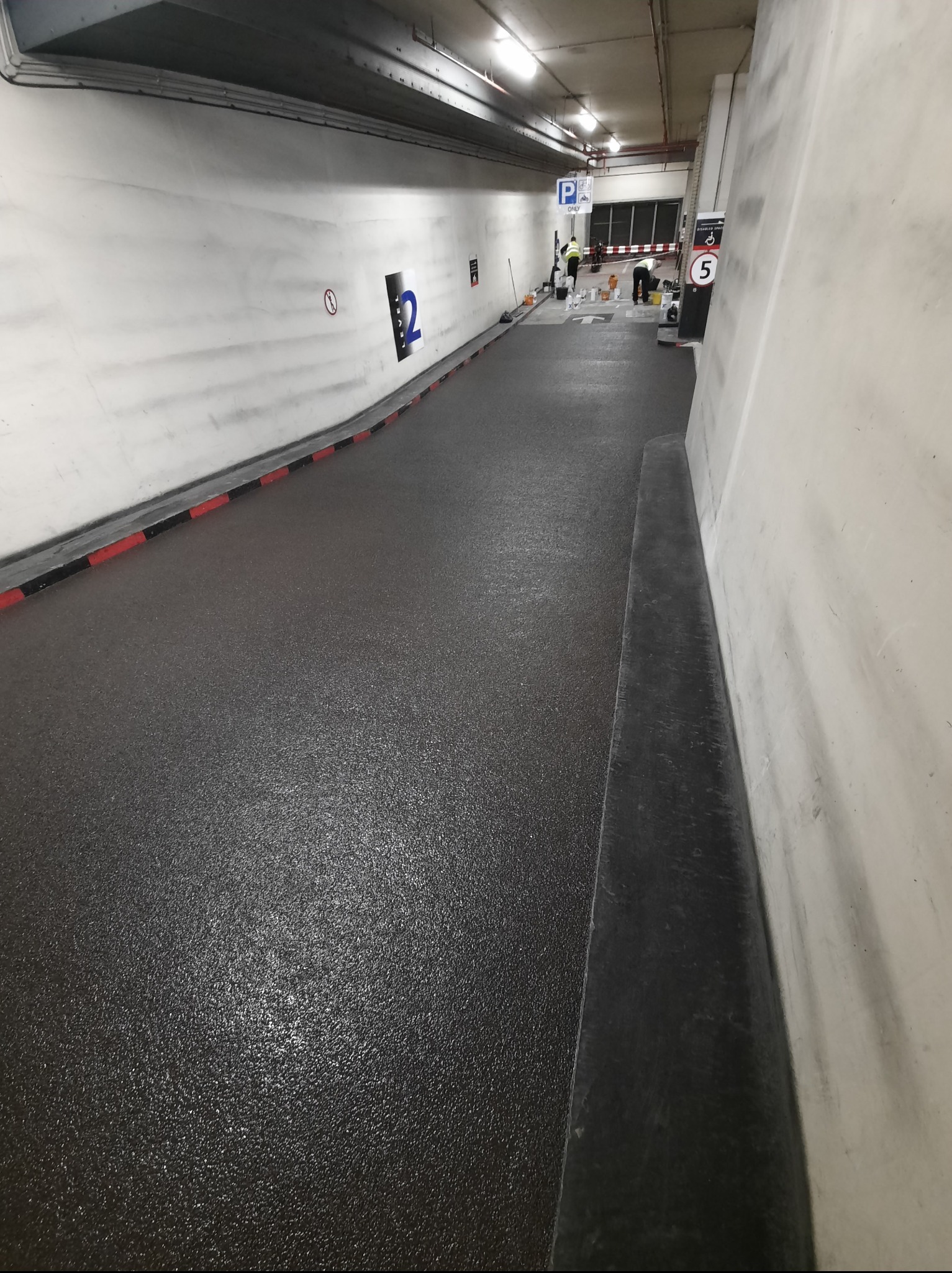 Prace wykończeniowe na parkingu podziemnym: świeżo pomalowana, błyszcząca posadzka w odcieniach szarości. W tle pracownicy i sprzęt.