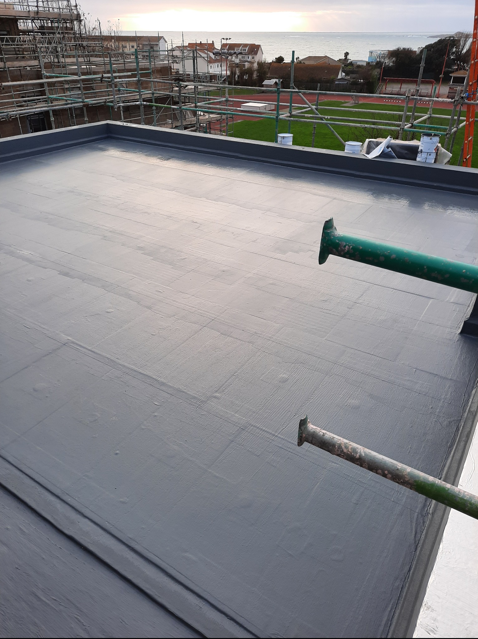 Szary, płaski dach po renowacji z widocznymi śladami wałka, z zielonymi podpórkami rusztowania i widokiem na morze w tle. Widoczne puszki z farbą.
