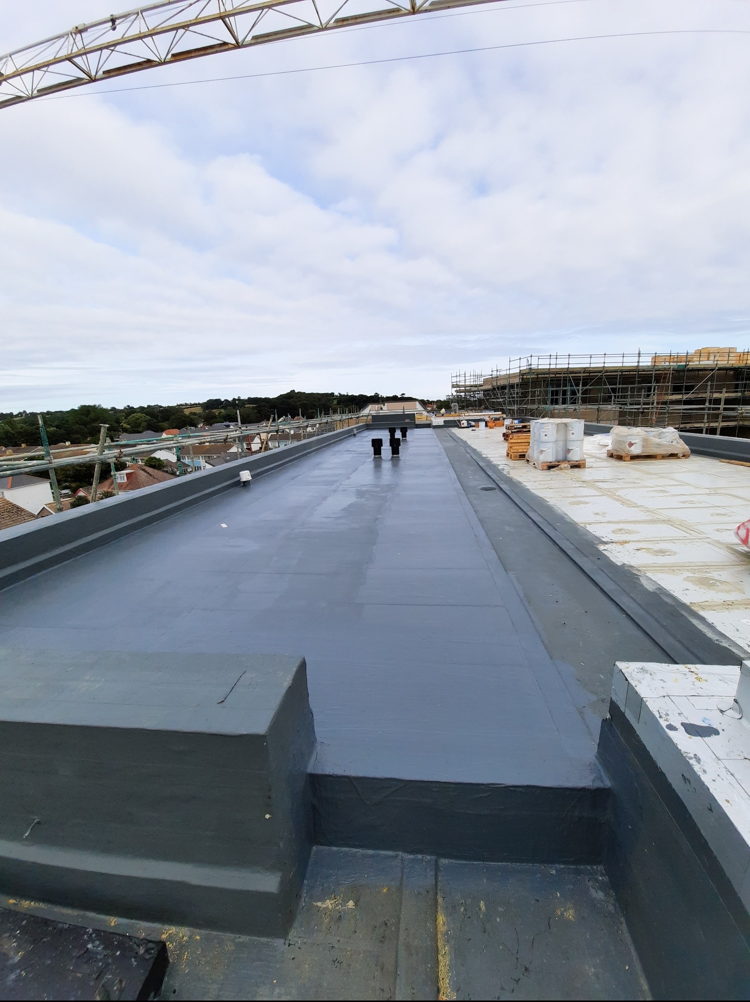 Szary, płaski dach po hydroizolacji z widocznymi kominami wentylacyjnymi i rusztowaniami w tle. Świeżo położona powłoka, brak detali wykończeniowych.