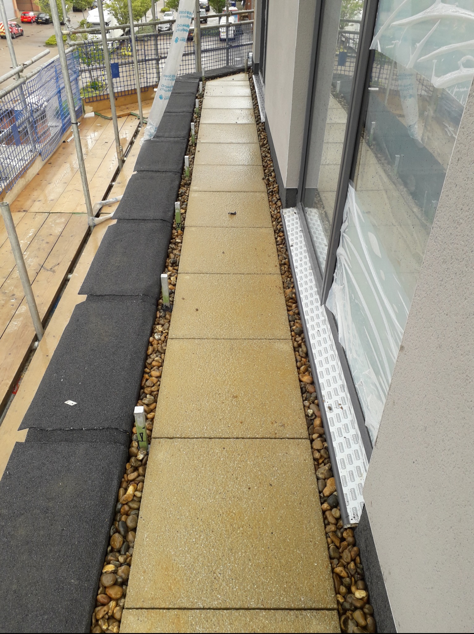 Wykończony taras na dachu z szarymi płytami gumowymi, jasnymi płytami chodnikowymi i kamieniami ozdobnymi, zabezpieczony rusztowaniem i folią ochronną.