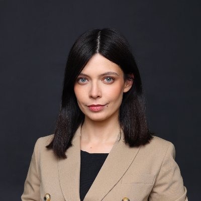 Portret kobiety z ciemnymi włosami i jasnymi oczami, ubrana w beżową marynarkę na tle ciemnoszarego tła. Delikatny makijaż, profesjonalny wygląd.