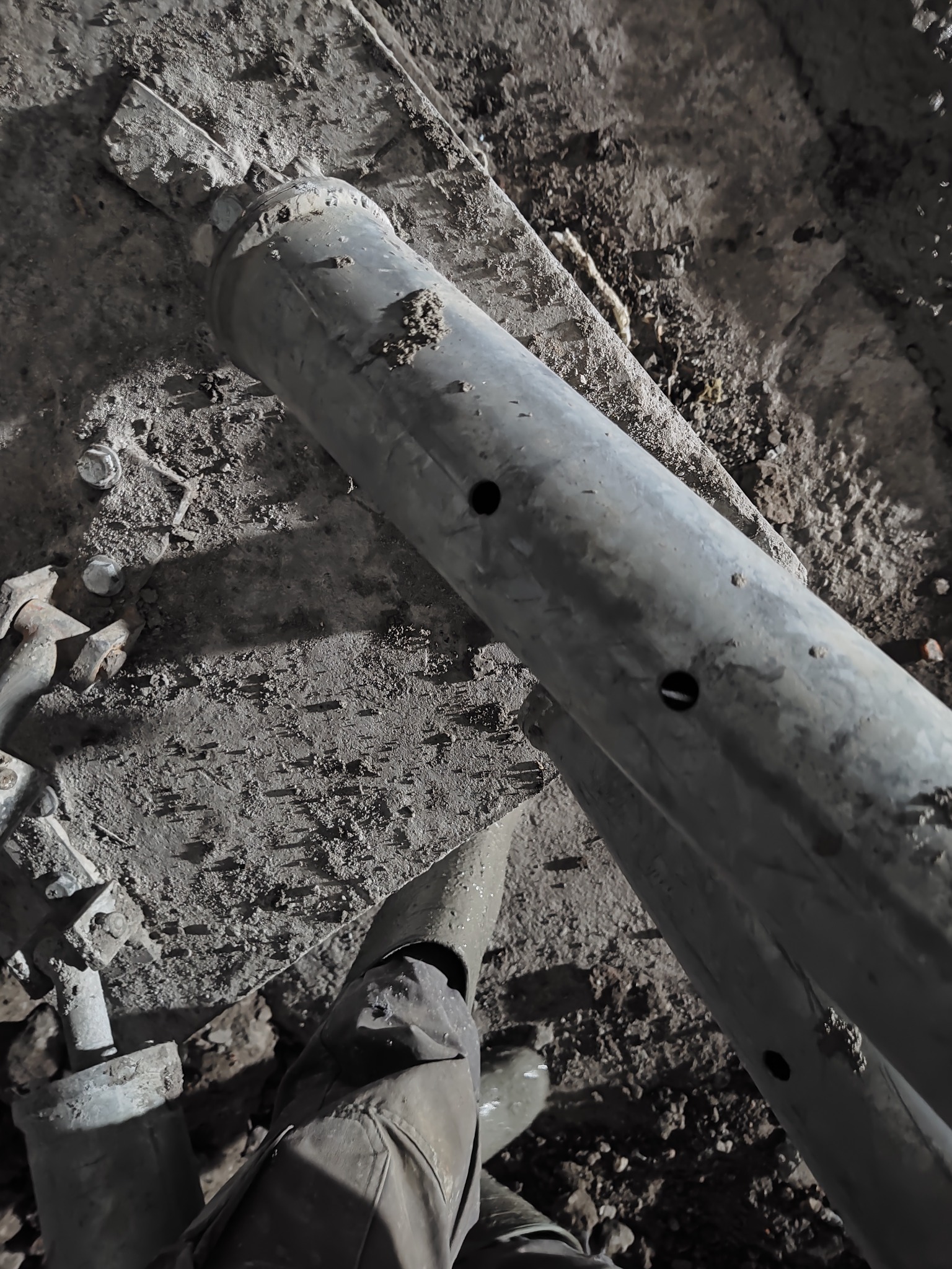 Szara, zbrojeniowa rura stalowa z otworami leży na ziemi z gruzem. Widoczne fragmenty butów i ubrania osoby, która prawdopodobnie układa rurę.