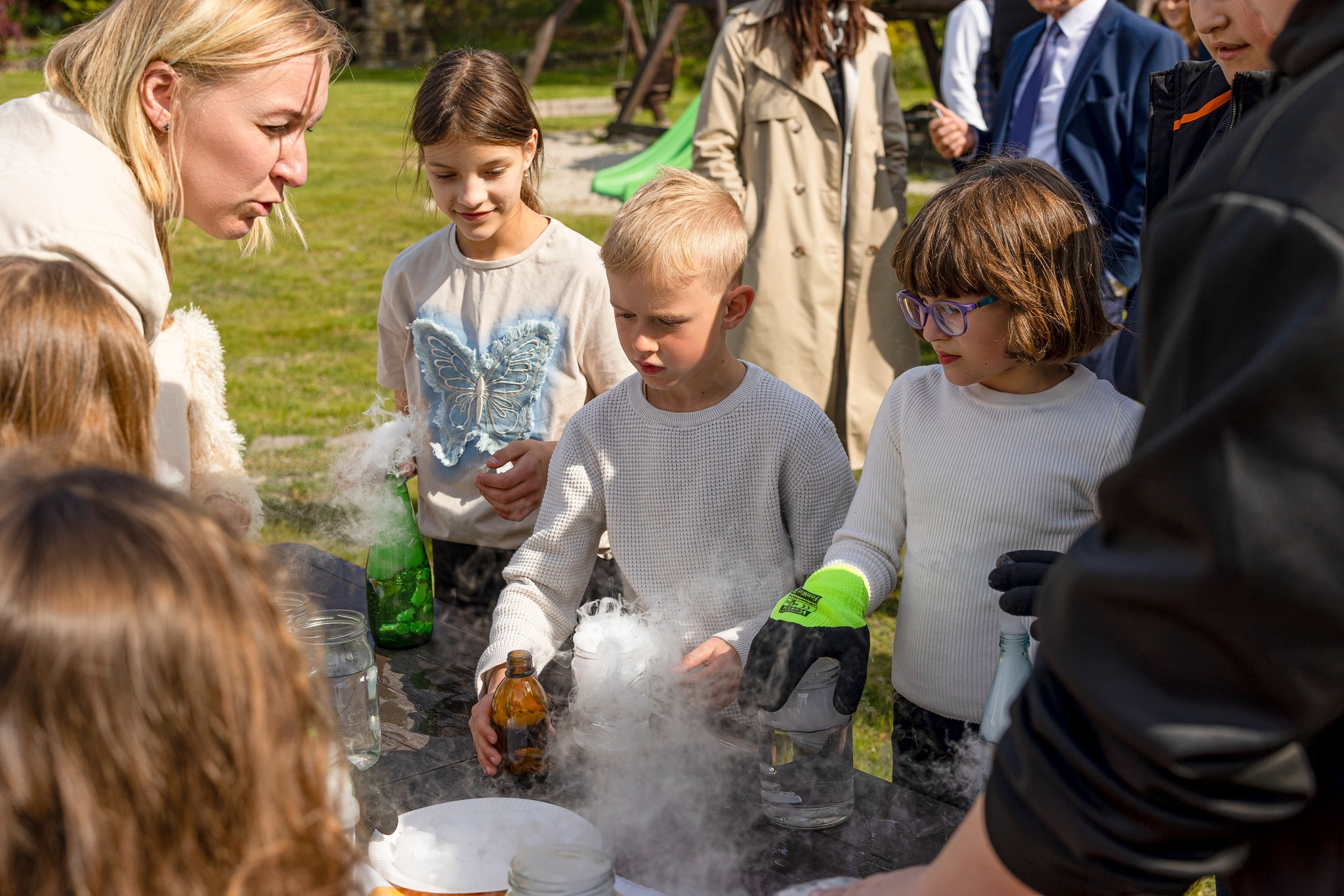 Dzieci obserwują eksperyment z suchym lodem prowadzony przez animatorkę. Widoczne szklane naczynia i unosząca się para. Uczestnicy w różnym wieku, na zewnątrz.