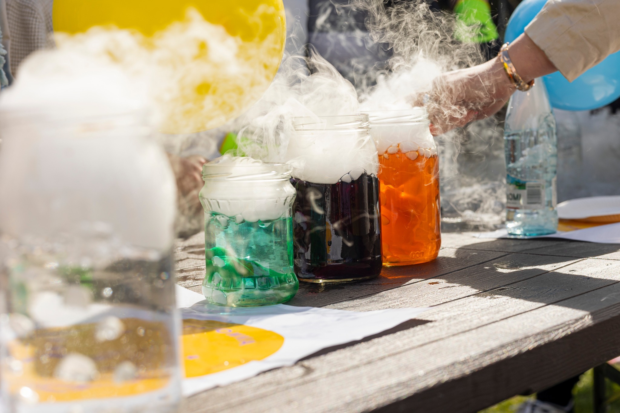Eksperymenty z suchym lodem w słoikach: kolorowe płyny, unosząca się para, balony w tle, ręka w mankiecie i bransoletce. Atrakcja na imprezę.