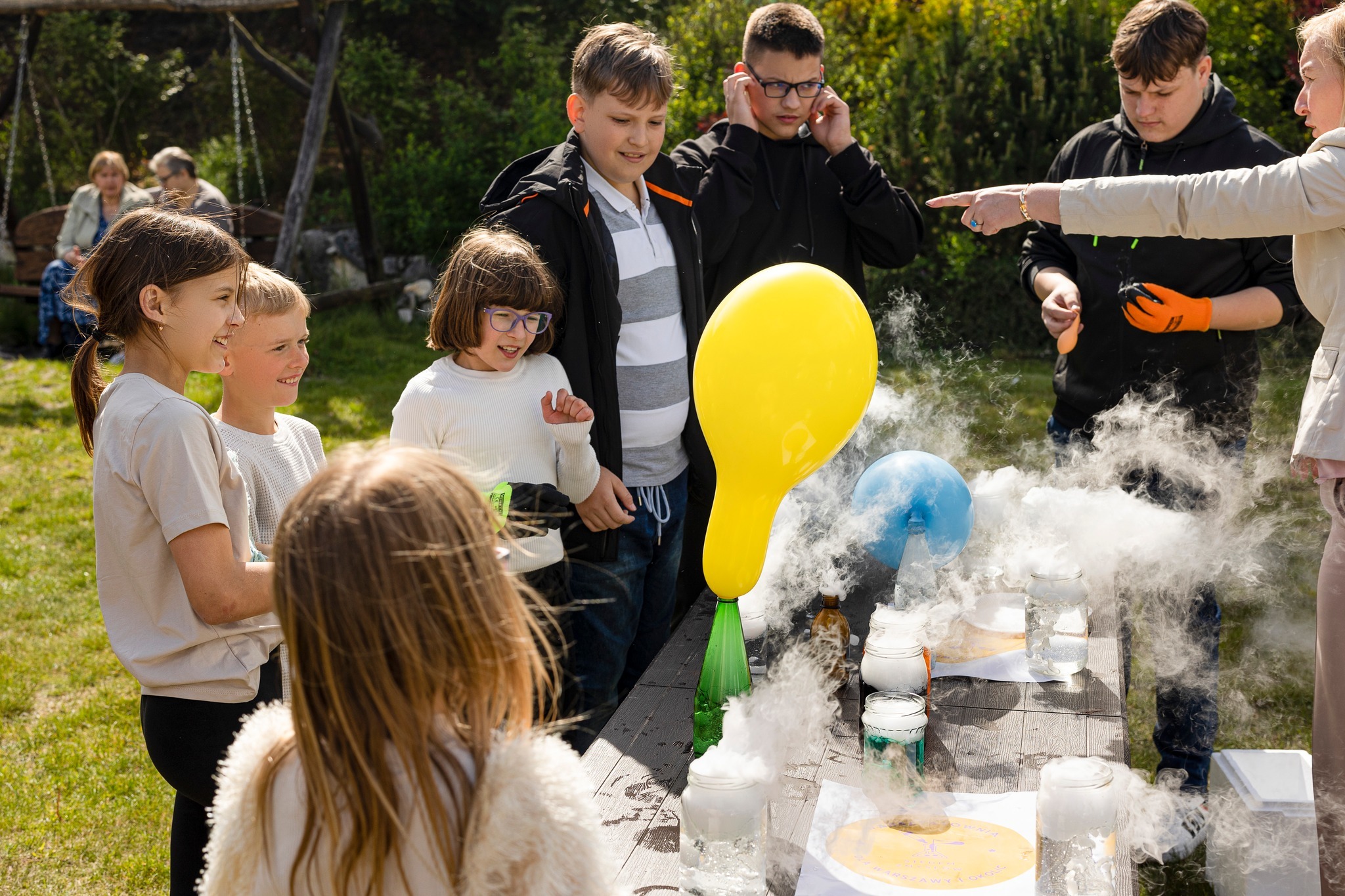Dzieci obserwują eksperyment naukowy z suchym lodem i balonami podczas imprezy z animatorem. Na stole słoiki i butelki z dymem.