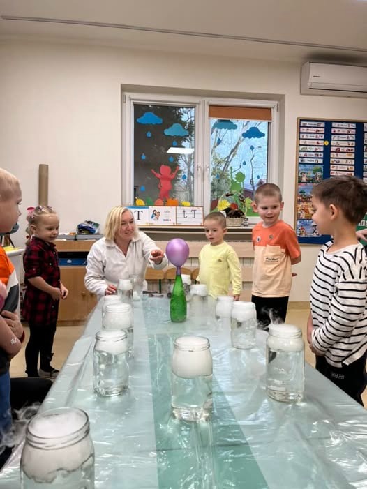Nauczycielka w białym fartuchu demonstruje eksperyment z dymiącymi słoikami i balonem grupie dzieci w jasnym pomieszczeniu z dekoracjami okiennymi.