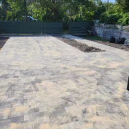 Ułożona kostka brukowa na podjeździe z miejscem na zieleń, widoczne ogrodzenie panelowe i betonowe. Kostka posypana piaskiem. Słoneczny dzień.