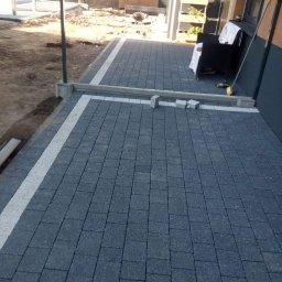 Dariusz sikorski - Ułożona, szara kostka brukowa z białym obramowaniem przy budynku. Fragment niedokończonego ogrodzenia w tle. Widoczne elementy wyposażenia ogrodu.