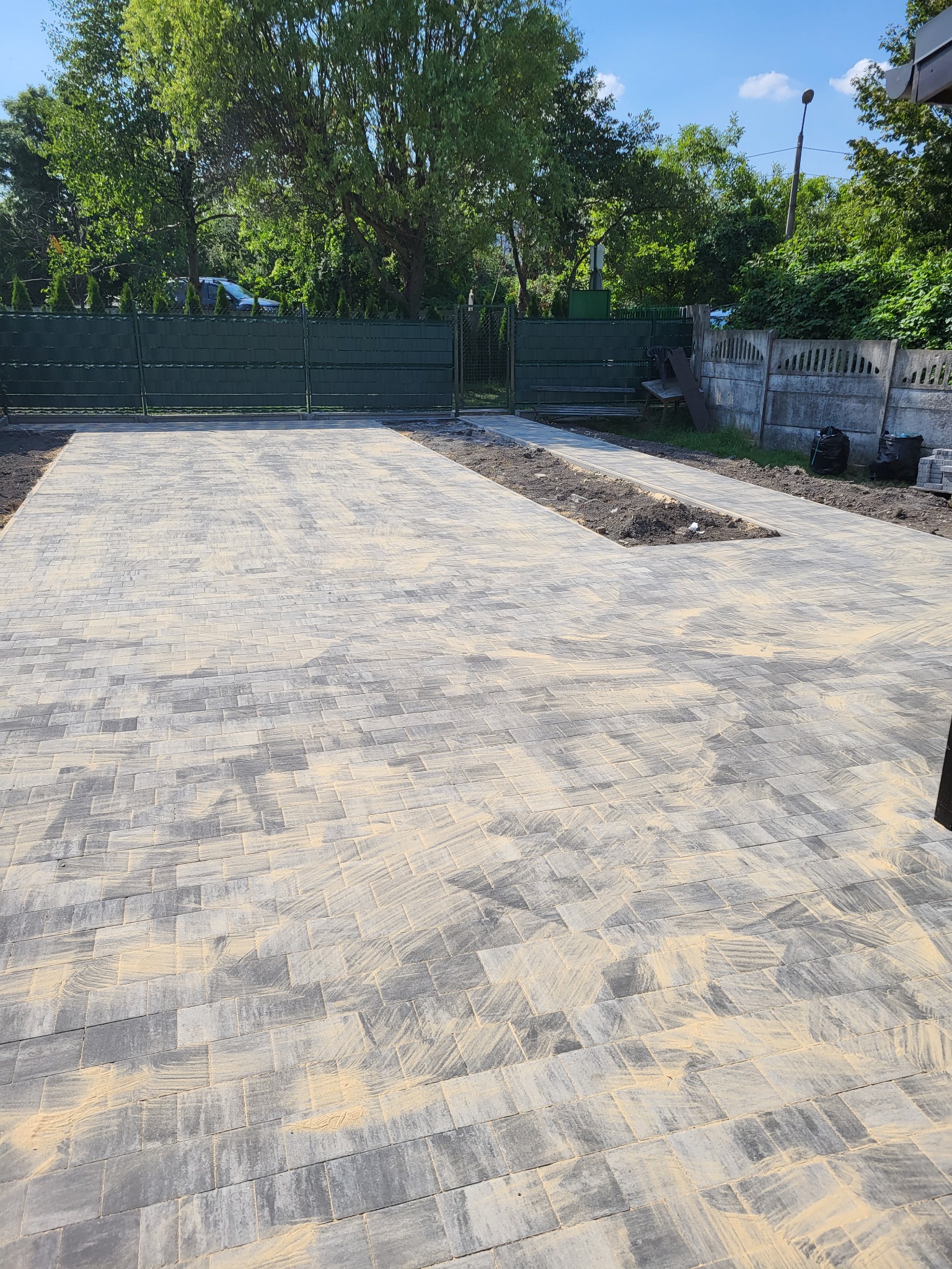 Ułożona kostka brukowa na podjeździe z miejscem na zieleń, widoczne ogrodzenie panelowe i betonowe. Kostka posypana piaskiem. Słoneczny dzień.