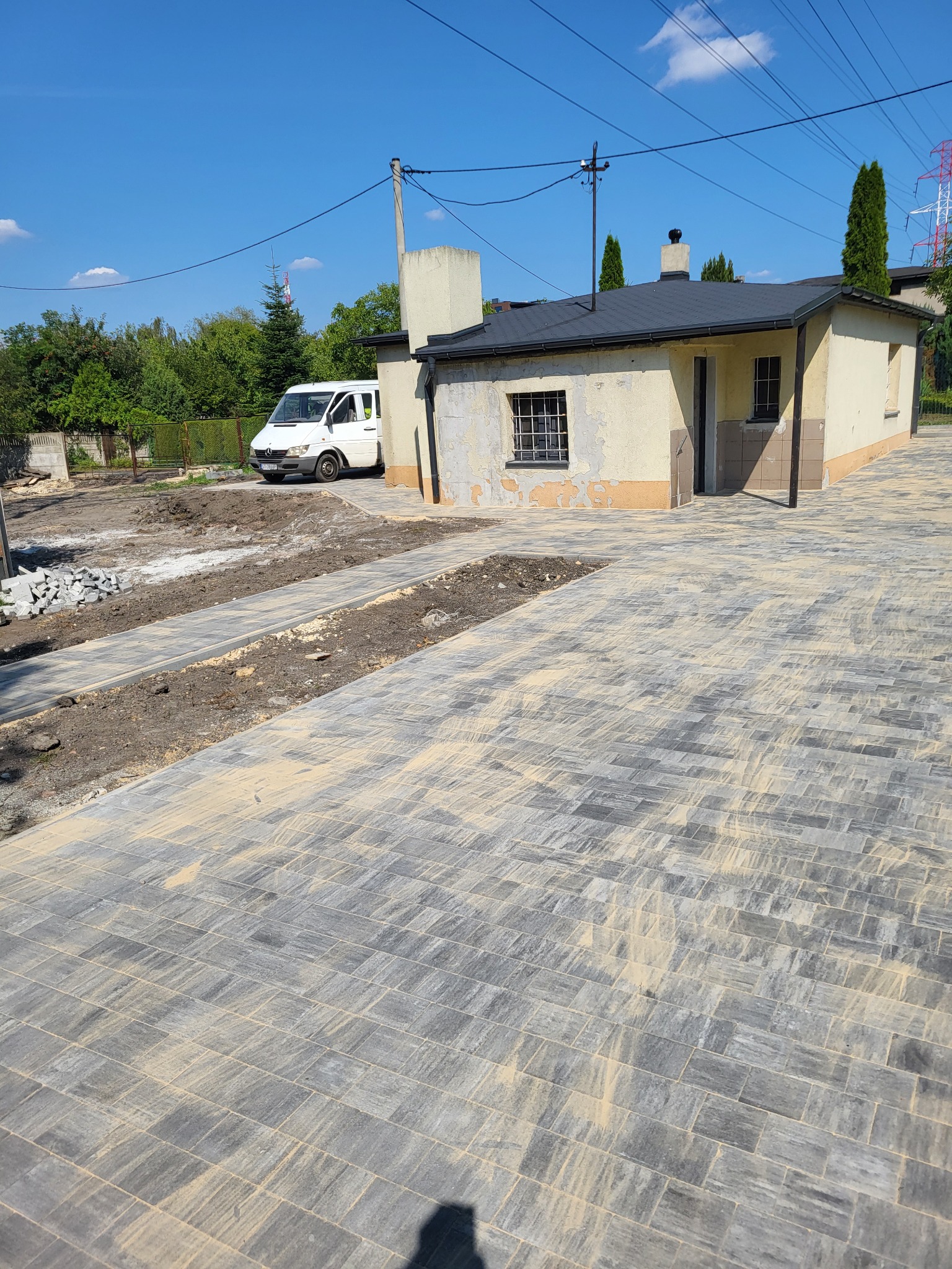 Układanie szarej kostki brukowej wokół budynku z białym vanem w tle. Widoczny fragment terenu przed budynkiem w trakcie przygotowania pod dalsze prace brukarskie.