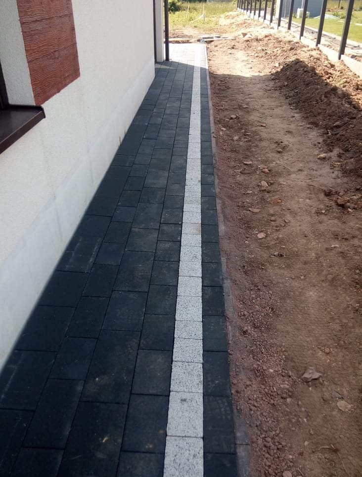 Ścieżka z ciemnej kostki brukowej z jasnym pasem pośrodku, biegnąca wzdłuż ściany budynku. Obok widoczne przygotowane podłoże.