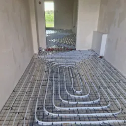 Białe rury ogrzewania podłogowego marki Keller ułożone na czarnej siatce w surowym wnętrzu, widok z góry, rozdzielacz na ścianie.