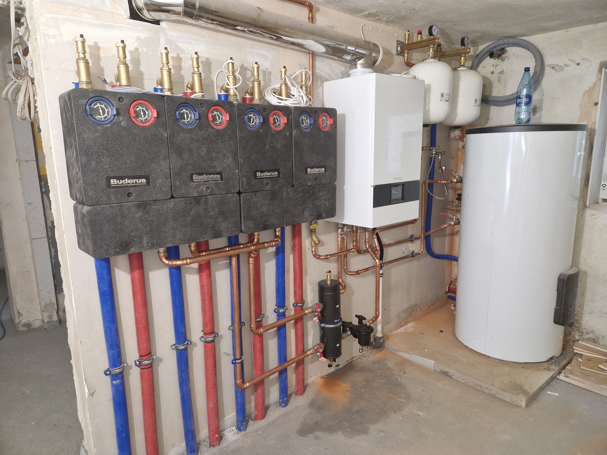 Zestaw kotłowni: piec gazowy, zasobnik CWU, naczynia wzbiorcze, rozdzielacz z grupami pompowymi. Miedziana instalacja, rury w izolacji czerwonej i niebieskiej.