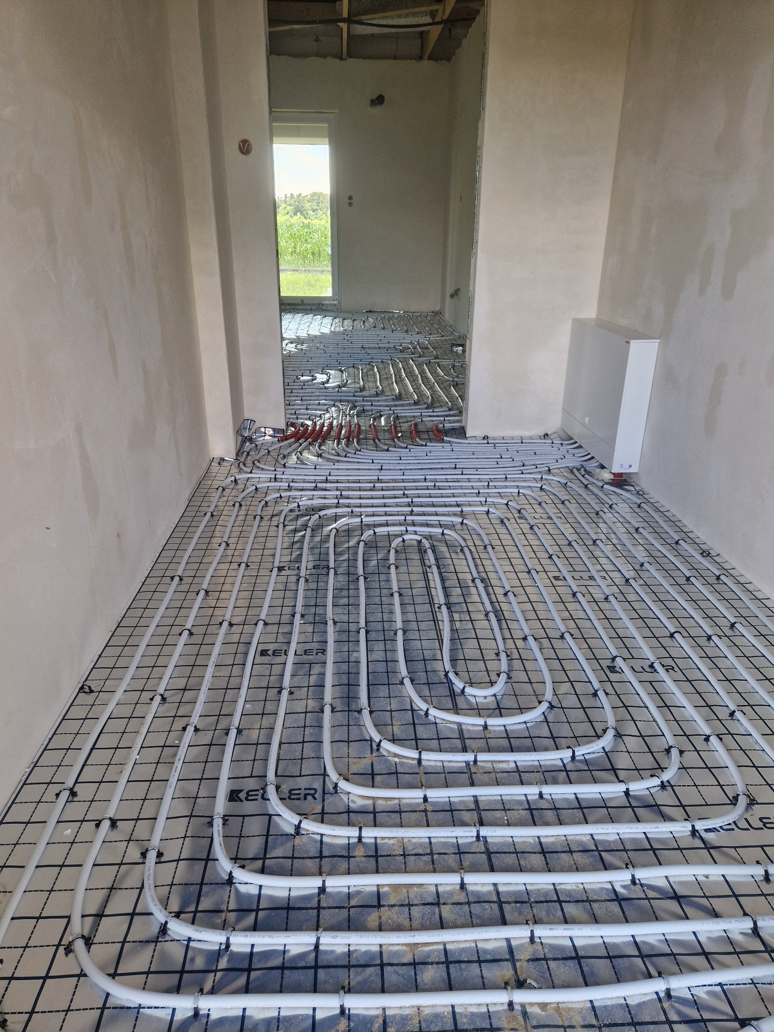 Białe rury ogrzewania podłogowego marki Keller ułożone na czarnej siatce w surowym wnętrzu, widok z góry, rozdzielacz na ścianie.