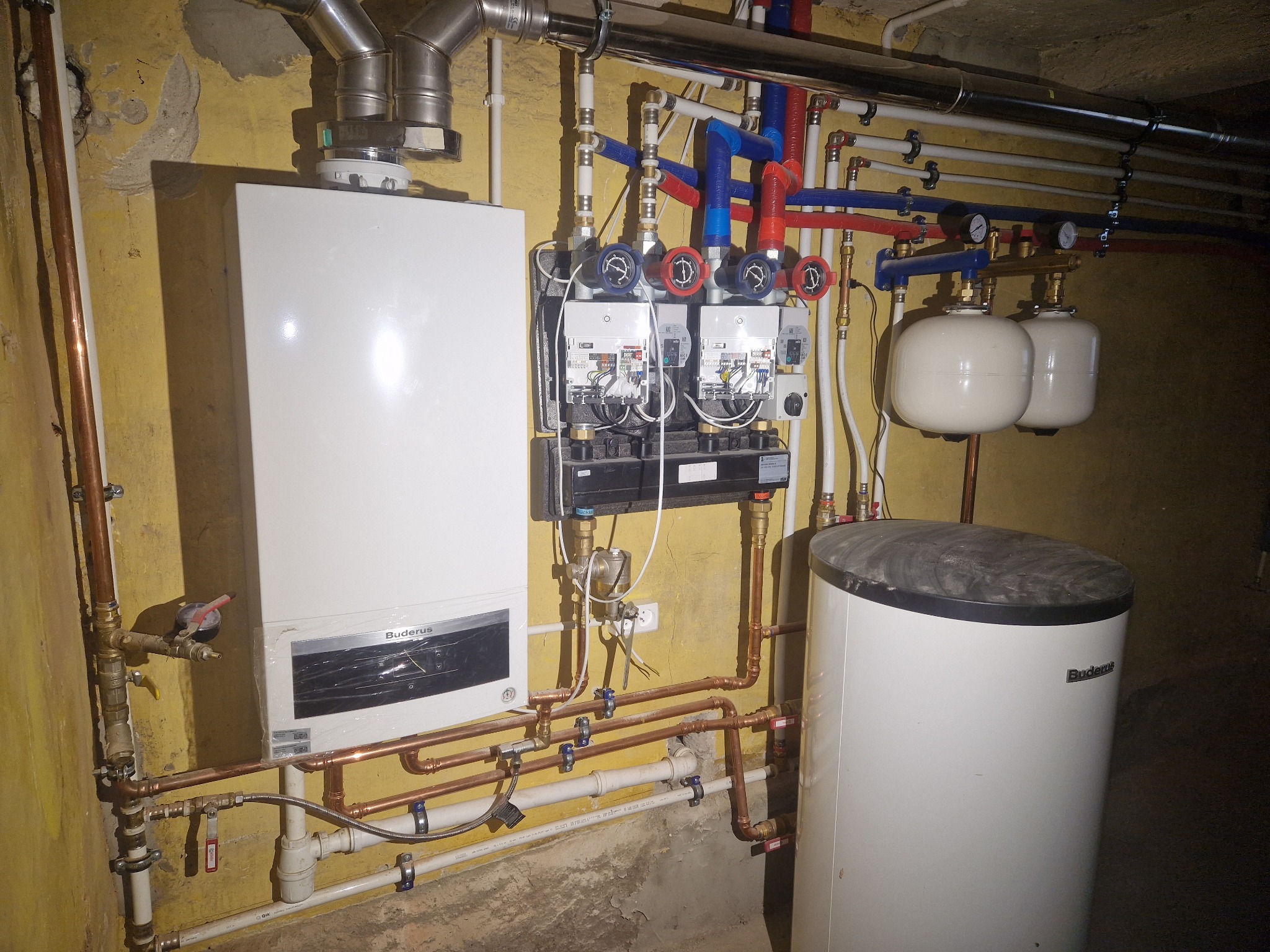 Złożona instalacja grzewcza w kotłowni: piec gazowy Buderus, zasobnik CWU, rozdzielacze, pompy obiegowe i rury miedziane na tle żółtej ściany piwnicy.