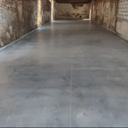 Gładka, szara posadzka przemysłowa w surowym wnętrzu z ceglanymi ścianami, wykończona białą listwą przypodłogową, widok na całe pomieszczenie.