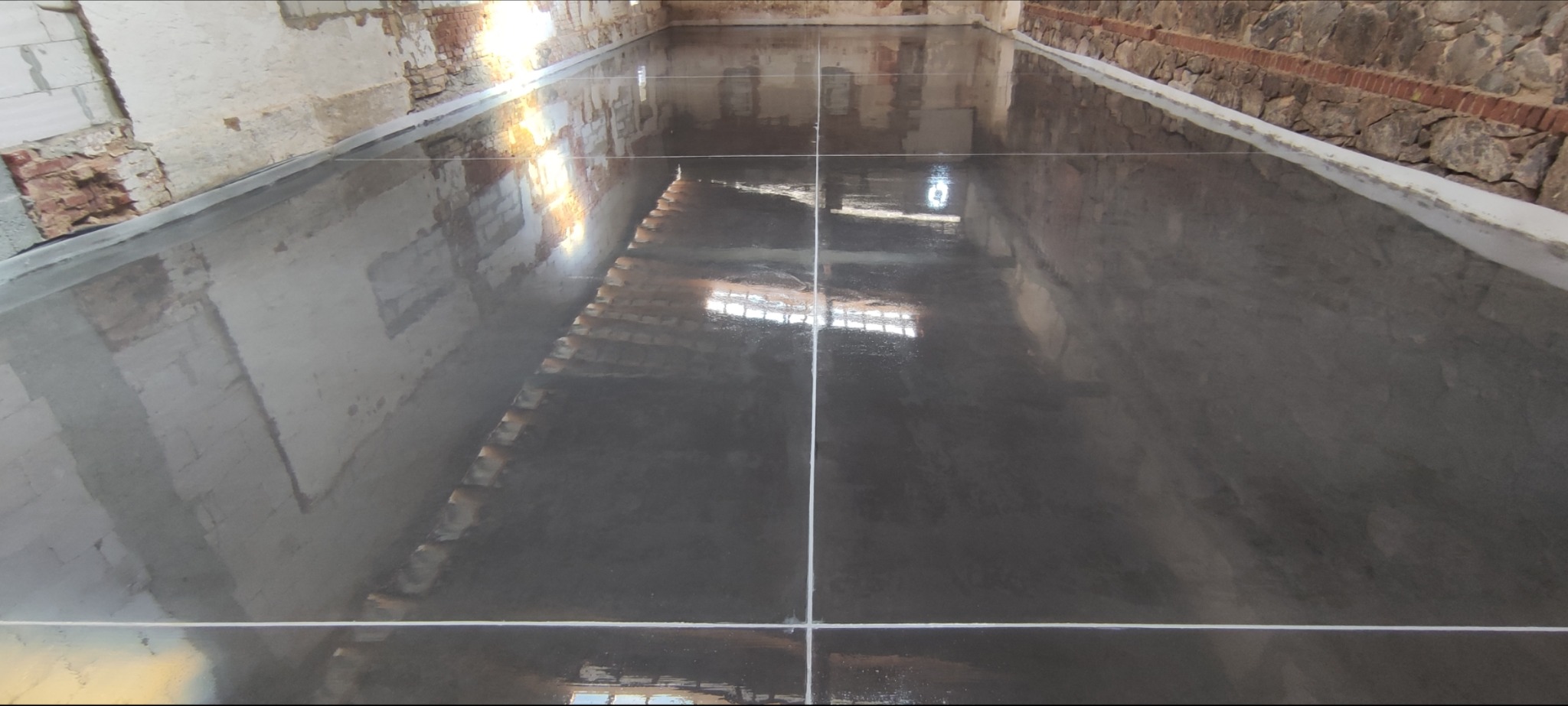 Świeżo wylana, gładka, szara posadzka przemysłowa w remontowanym wnętrzu z surowymi, ceglanymi ścianami i widocznymi liniami podziału.