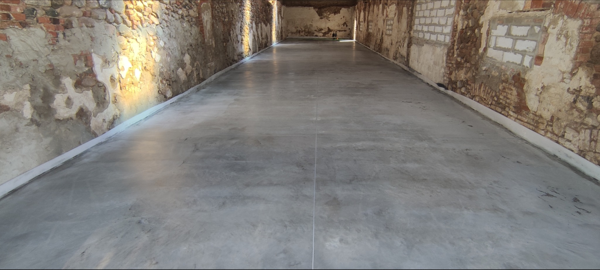 Gładka, szara posadzka przemysłowa w surowym wnętrzu z ceglanymi ścianami, wykończona białą listwą przypodłogową, widok na całe pomieszczenie.