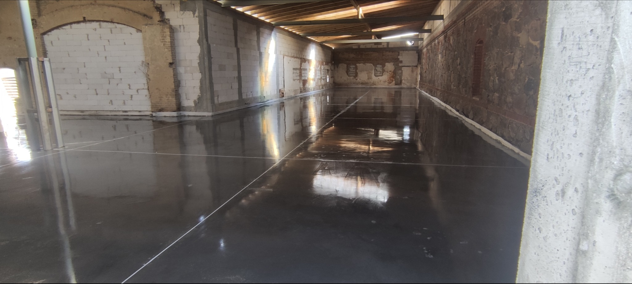 Błyszcząca, szara posadzka przemysłowa w surowym wnętrzu z ceglanymi i betonowymi ścianami. Widoczne linie dylatacyjne i refleksy światła.