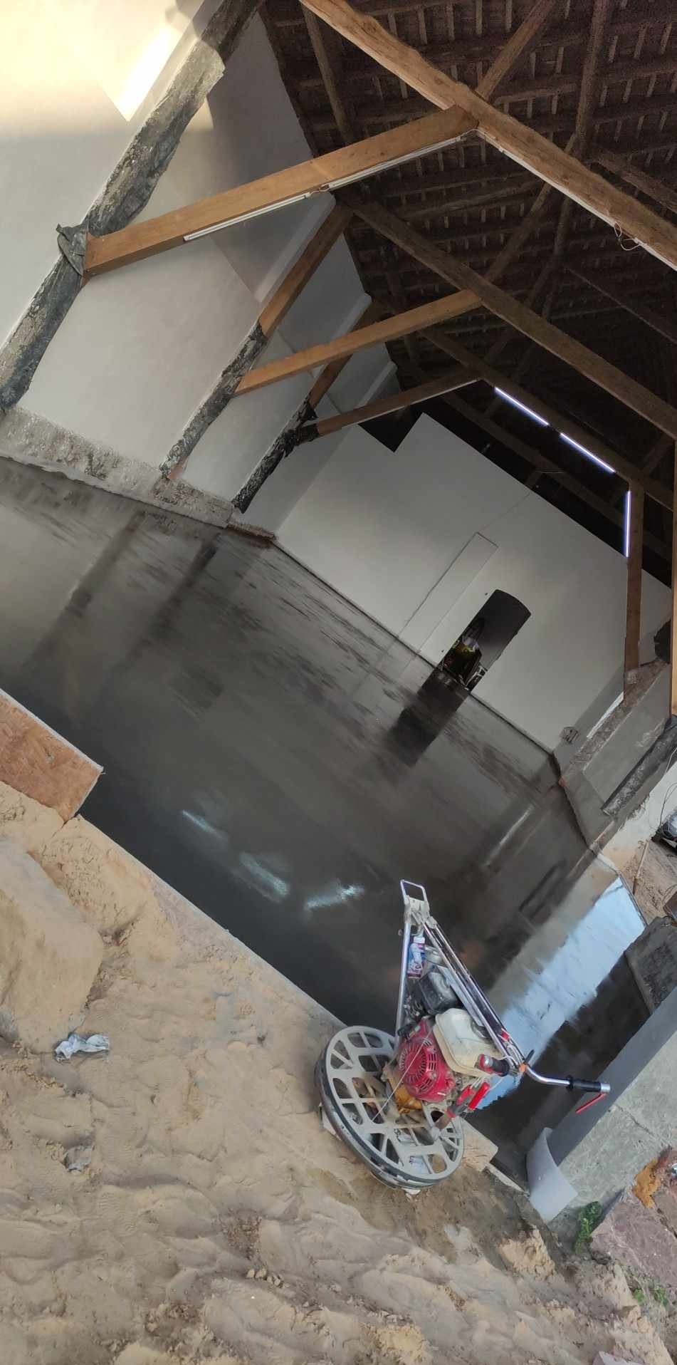 Wnętrze z surowym drewnianym stropem po wylaniu błyszczącej, ciemnej posadzki. Na pierwszym planie maszyna do polerowania. Widoczne ślady prac budowlanych.