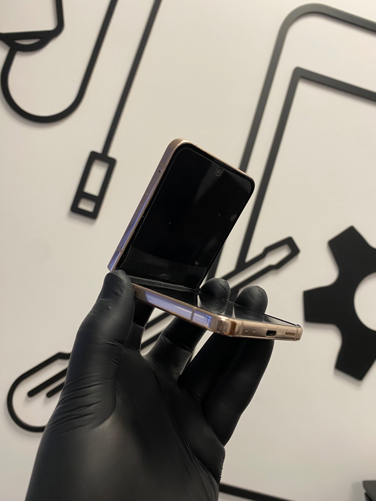 Złoty składany smartfon trzymany w dłoni w czarnej rękawiczce, na tle białej ściany z czarnymi, graficznymi elementami narzędzi serwisowych.