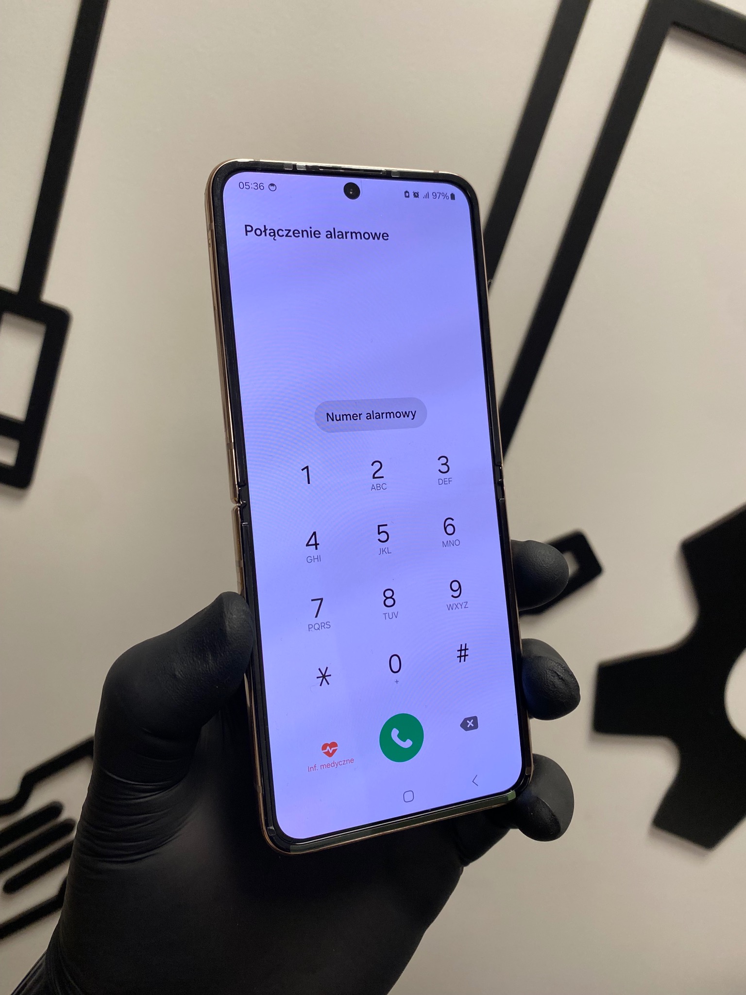 Składany smartfon z ekranem alarmowym trzymany w dłoni w czarnej rękawiczce, na tle białej ściany z czarnym wzorem. Widoczny interfejs wybierania numeru alarmowego.