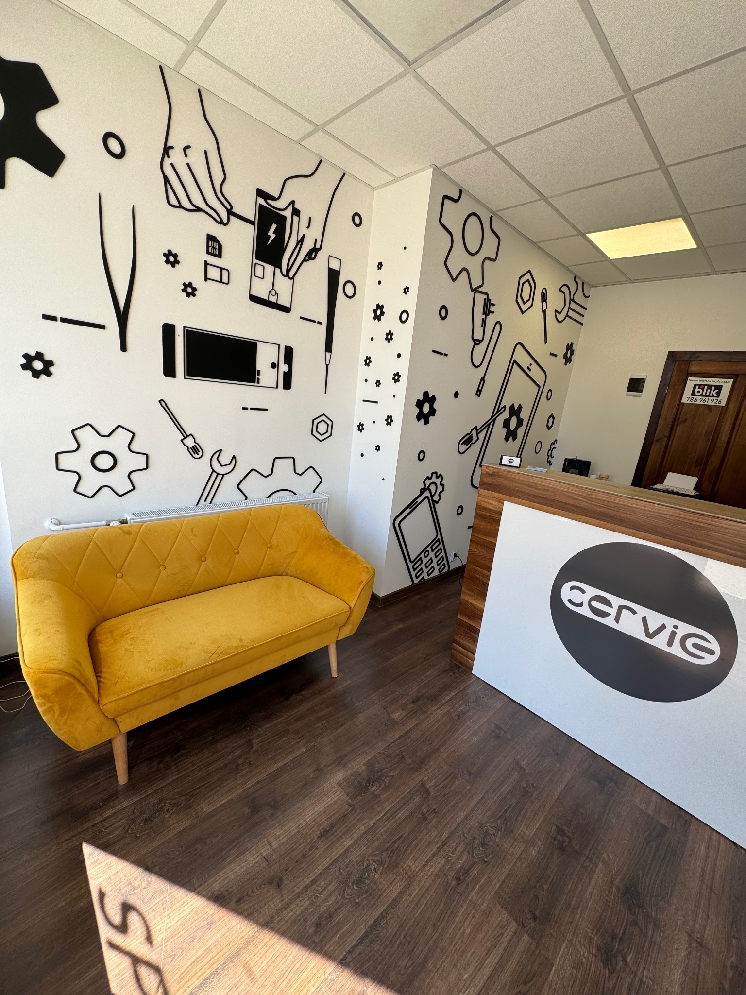 Wnętrze serwisu: żółta sofa, grafiki na ścianie z narzędziami i telefonami, lada z logo 'cervie', drewniana podłoga. Jasne oświetlenie.
