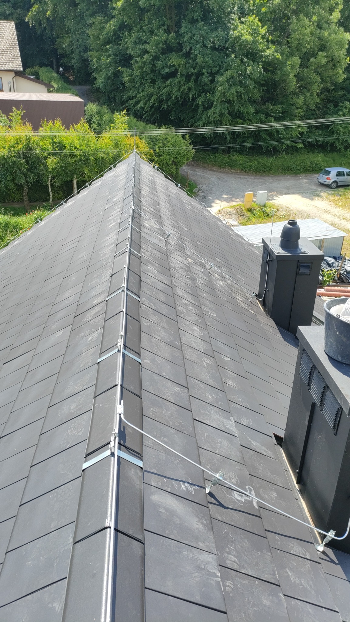 Szary dach z dachówki ceramicznej z zamontowaną instalacją odgromową, widok z góry, na tle zieleni i zabudowań.