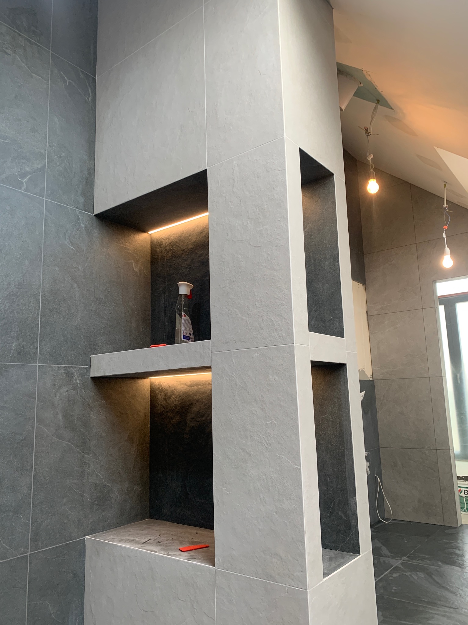 Szara łazienka w trakcie wykańczania: płytki imitujące kamień, wnęki z podświetleniem LED, widoczne niedokończone elementy sufitu i wiszące żarówki.