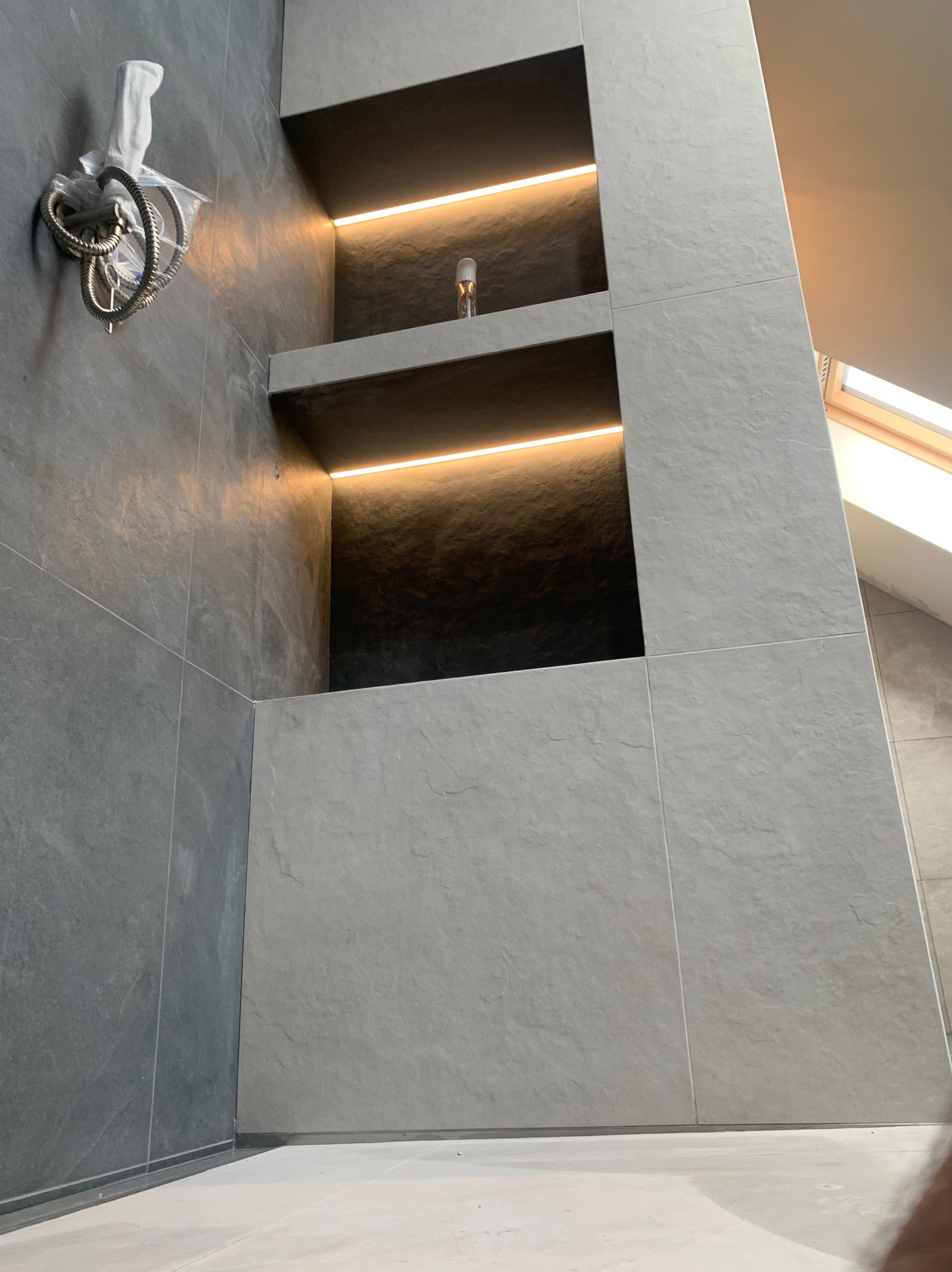 Nowoczesna łazienka z szarymi płytkami i podświetlanymi półkami wnękowymi. Niezamontowana słuchawka prysznicowa z wężem w folii. Okno dachowe w tle.