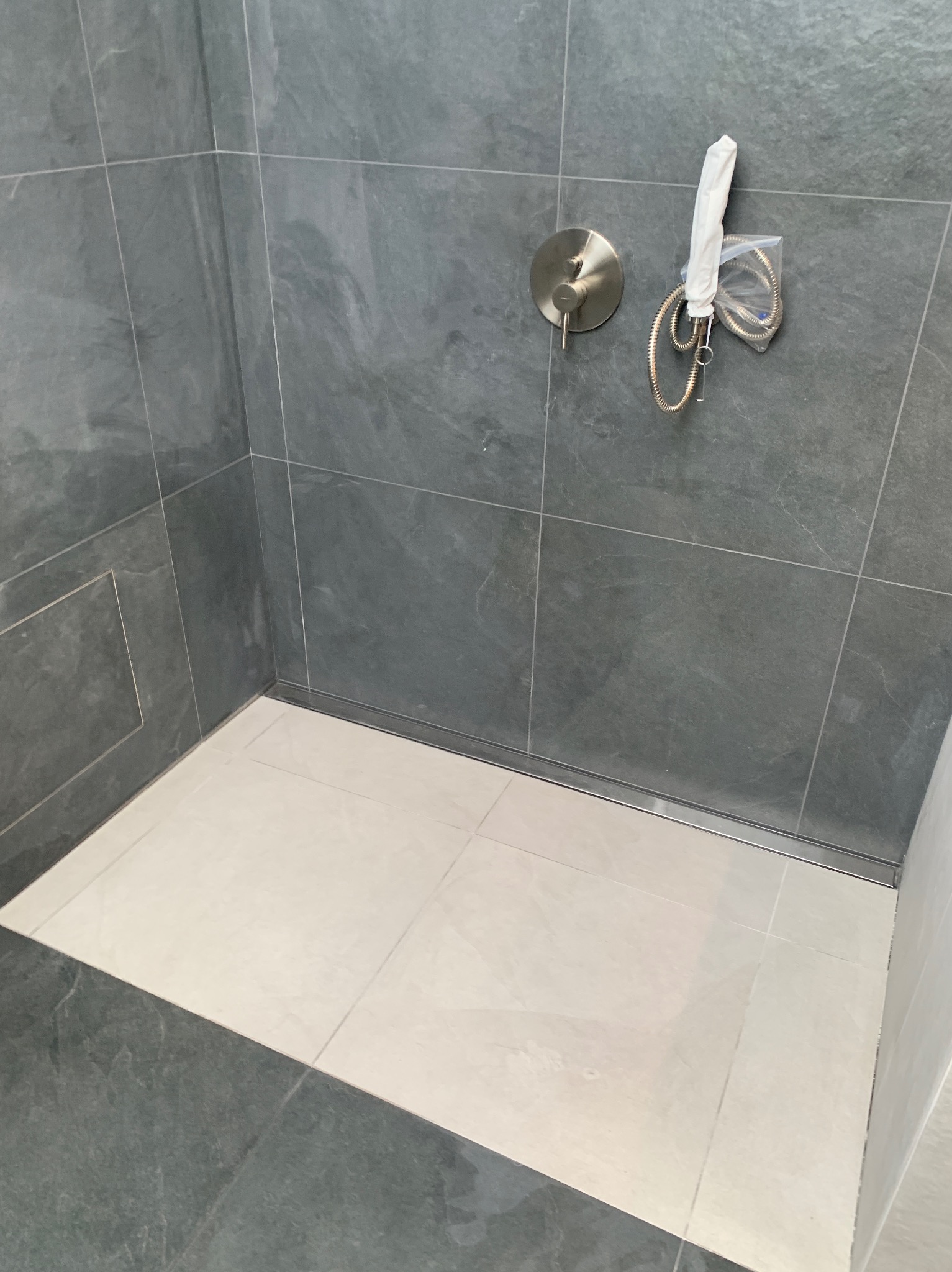 Wykończony brodzik z szarymi, teksturowanymi płytkami ściennymi i jasnymi płytkami podłogowymi. Widoczna armatura prysznicowa i odpływ liniowy.