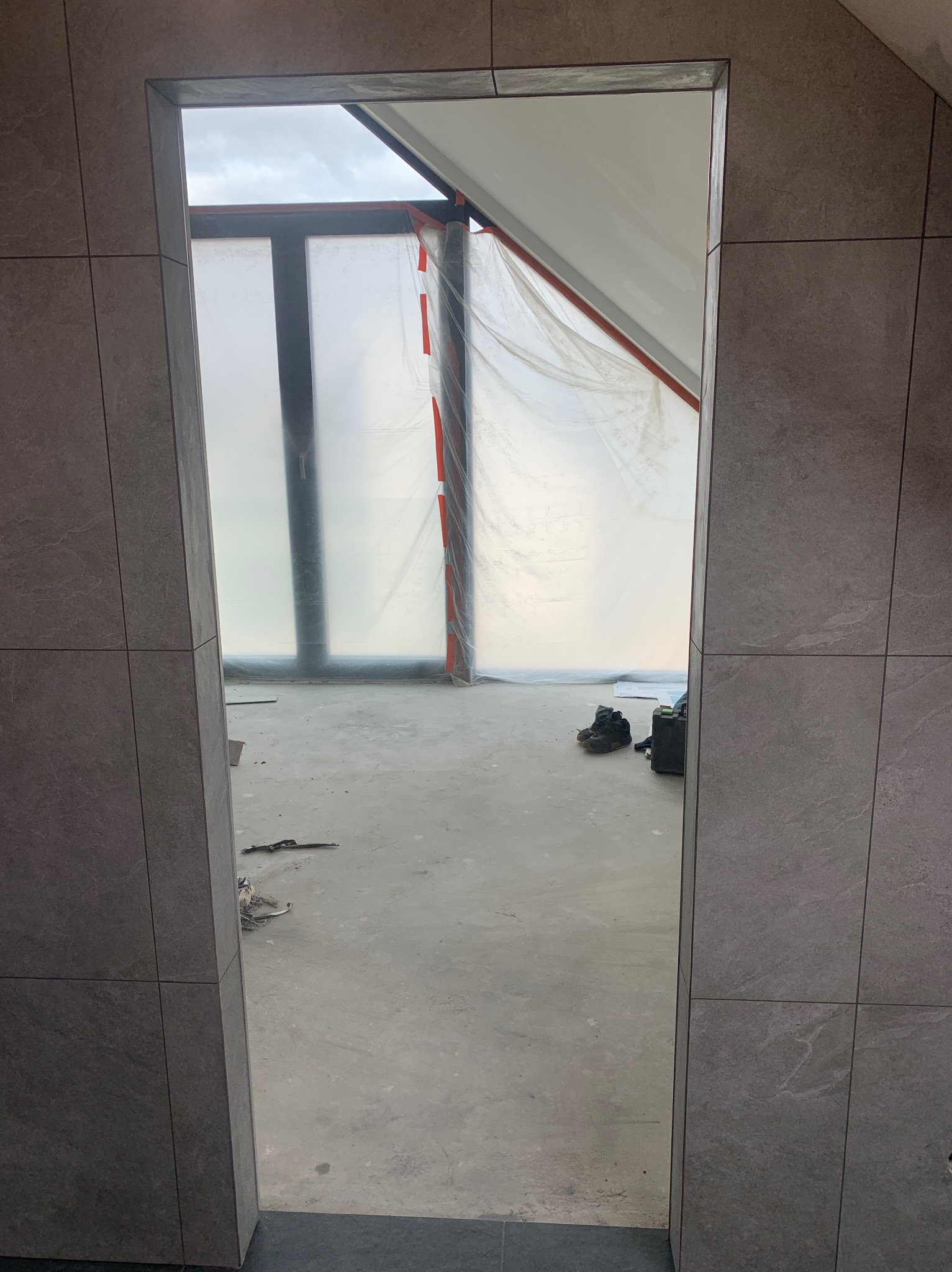 Wykończona wnęka drzwiowa z szarymi płytkami, widok na remontowane poddasze z oknami zabezpieczonymi folią. Widoczne narzędzia i buty.