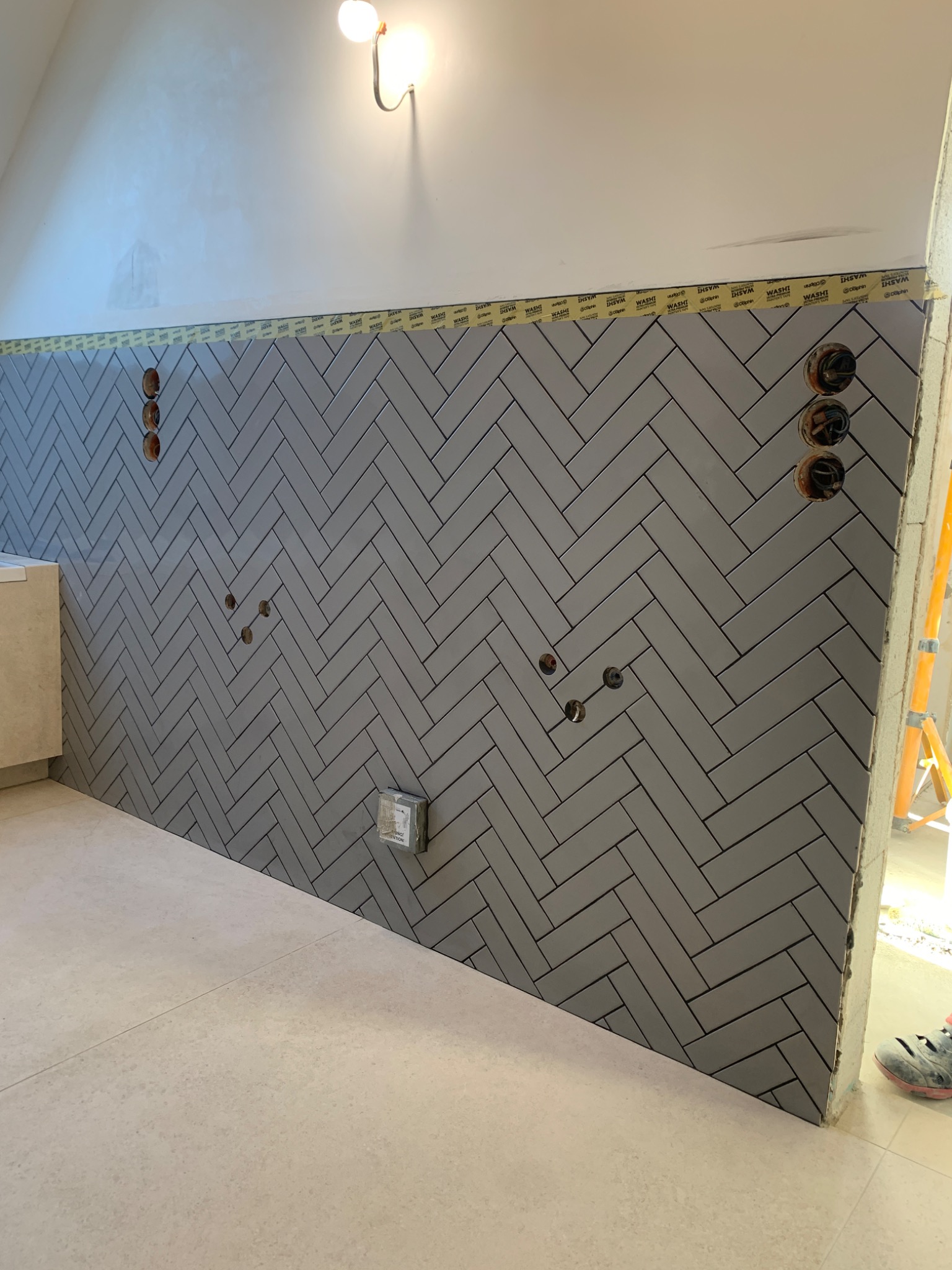 Ukończona ściana z szarych płytek ułożonych w jodełkę, zabezpieczona taśmą malarską. Widoczne puszki elektryczne i fragment podłogi z jasnych płytek.
