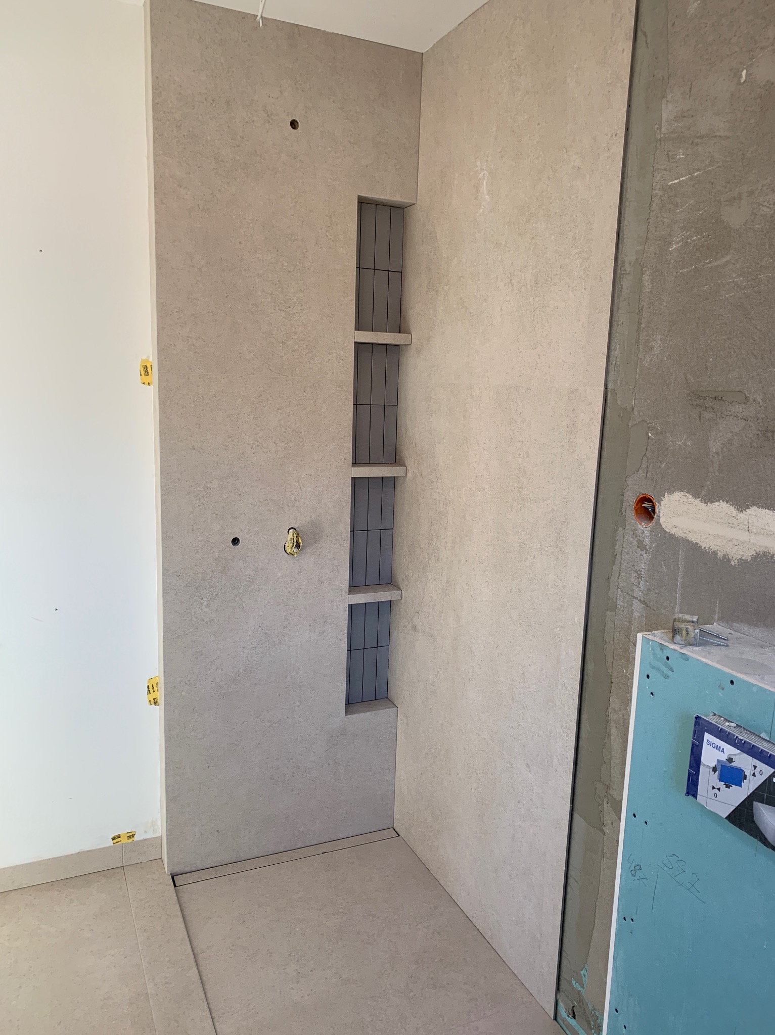 Wykończona wnęka z półkami w łazience, obłożona płytkami w odcieniach beżu i szarości. Widoczne narzędzia glazurnicze i płyta karton-gips, co wskazuje na etap prac wykończeniowych.