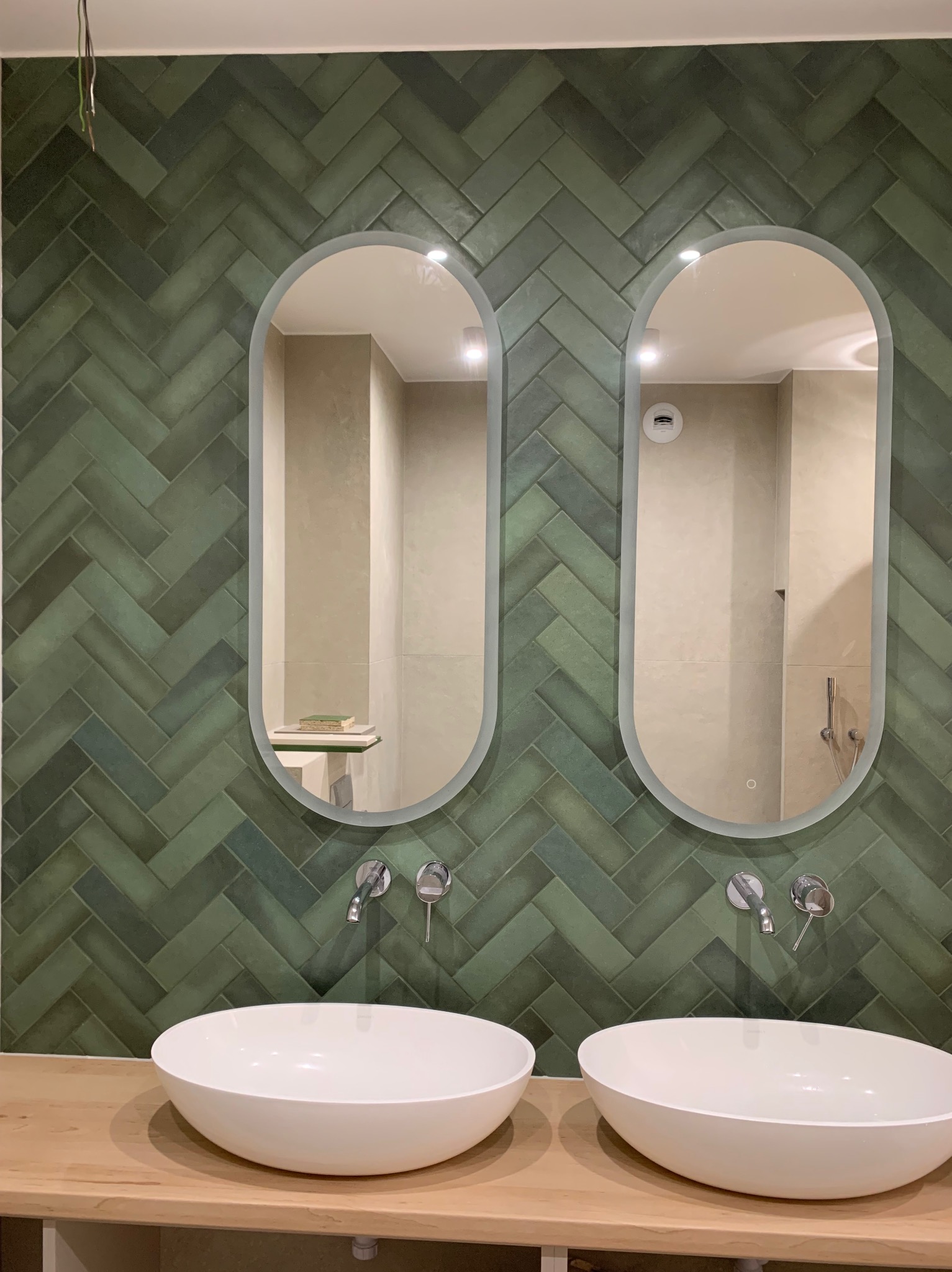 Wykończona łazienka z zieloną, ułożoną w jodełkę glazurą, owalnymi lustrami z podświetleniem LED i białymi, owalnymi umywalkami nablatowymi.