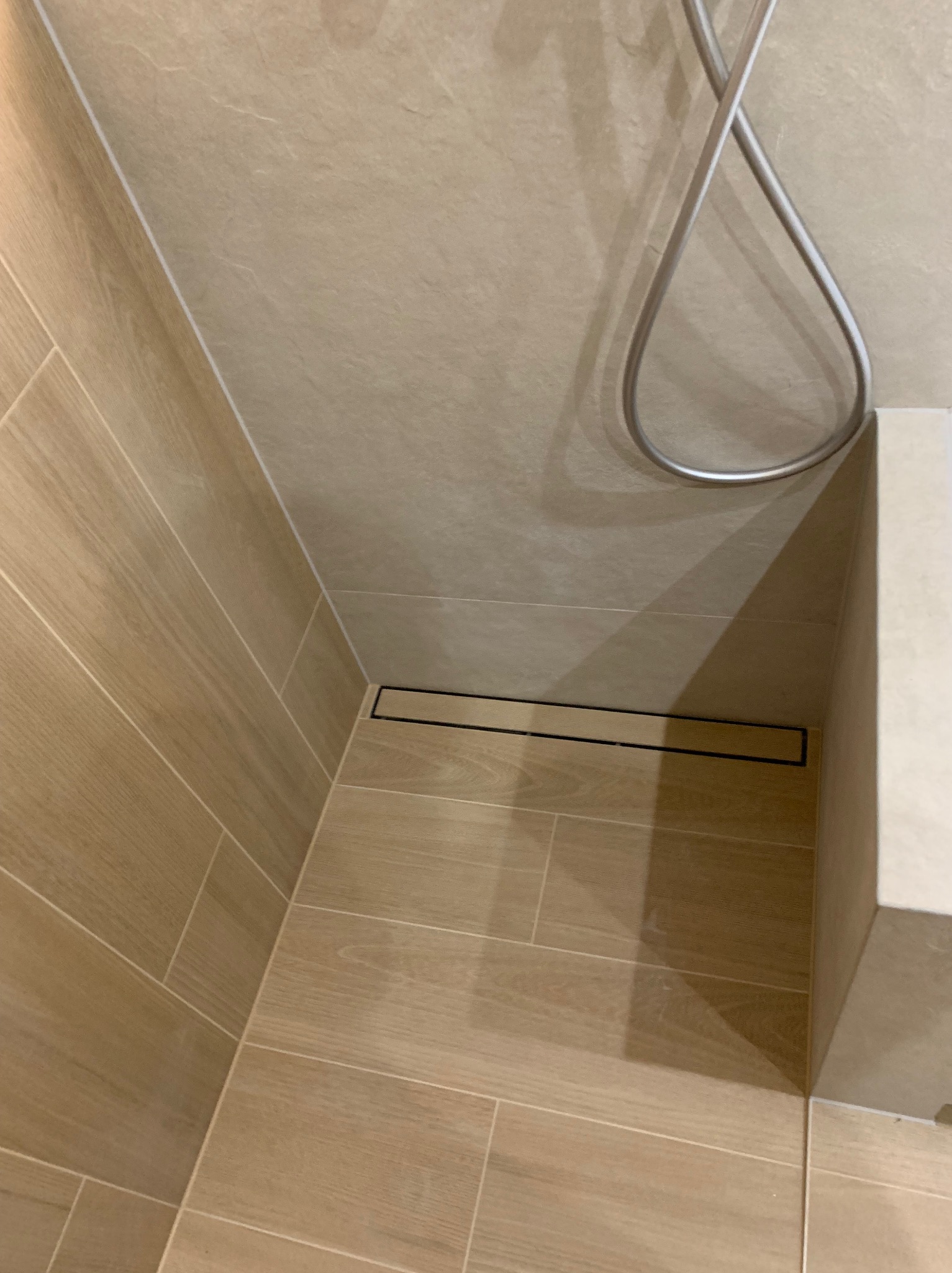 Beżowa łazienka z odpływem liniowym i chromowaną słuchawką prysznicową. Płytki imitujące drewno na ścianach i podłodze, minimalistyczny design.