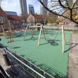 Nowoczesny plac zabaw z huśtawkami i drabinkami na zielonej nawierzchni, w tle budynki mieszkalne i wieżowce. Całość ujęta w słoneczny dzień.