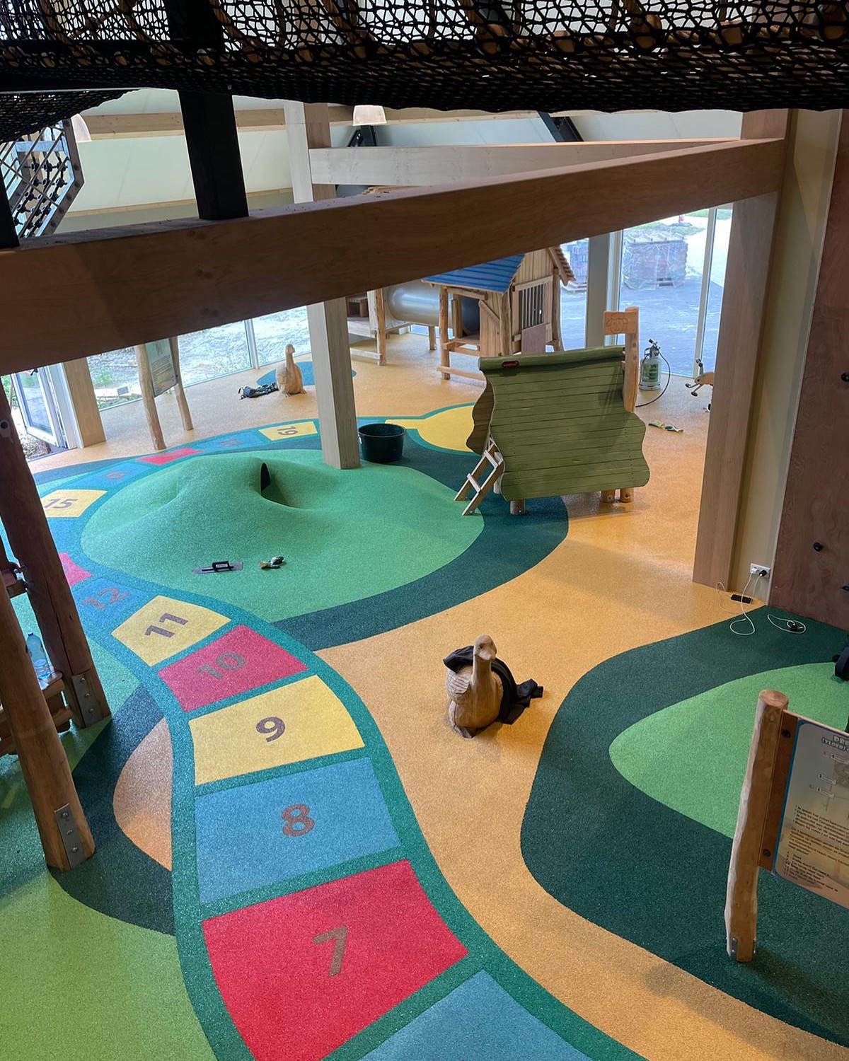 Kolorowy plac zabaw wewnątrz budynku z drewnianymi elementami i interaktywnymi grami podłogowymi, widok z góry.