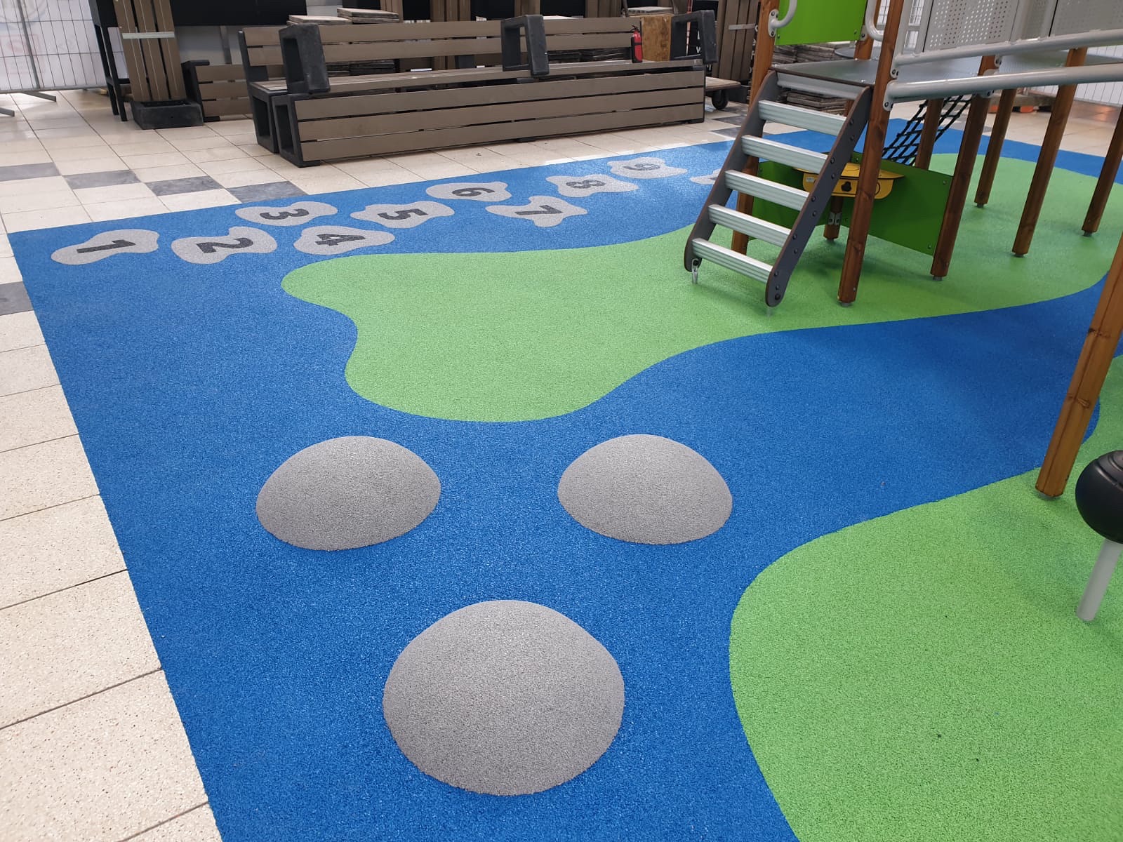 Nowoczesny plac zabaw z gumową nawierzchnią w kolorach niebieskim i zielonym, z elementami do skakania i drabinką.