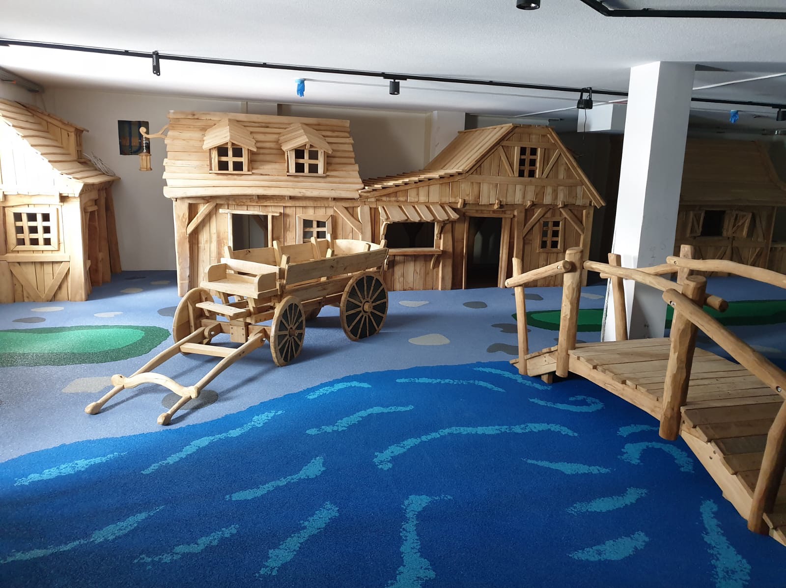 Wyjątkowy, drewniany plac zabaw w stylu wioski z wozem i mostkiem na niebieskiej podłodze imitującej rzekę. Realizacja z dbałością o detale.