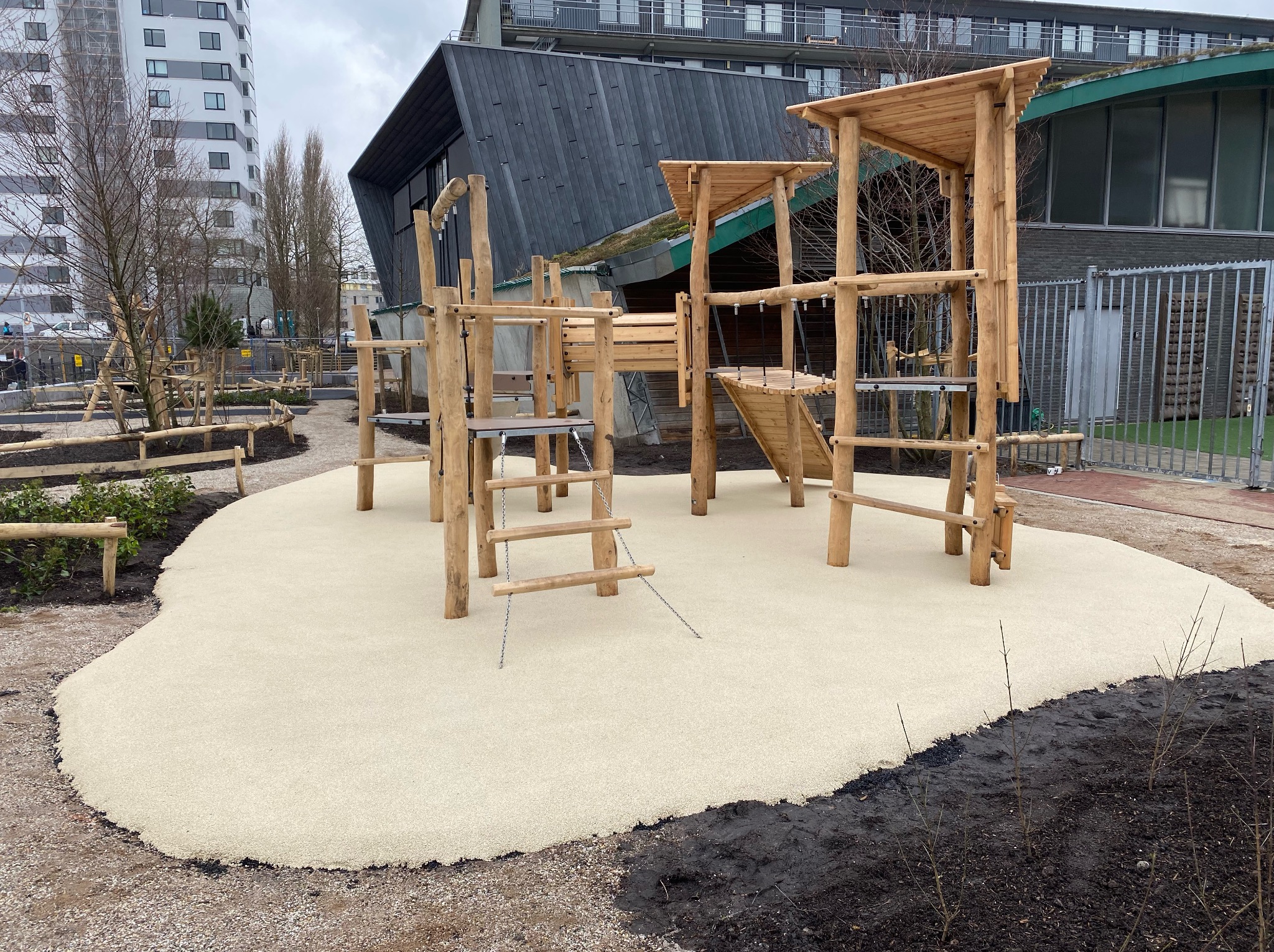 Drewniany plac zabaw z wieżami i mostkami na beżowej, bezpiecznej nawierzchni. W tle budynki mieszkalne i zielony dach. Nowoczesny design.