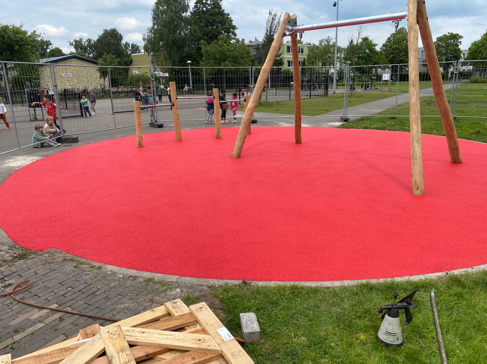 Nowy plac zabaw z czerwonym, bezpiecznym podłożem i drewnianymi konstrukcjami do wspinaczki, otoczony ogrodzeniem budowlanym. W tle dzieci.