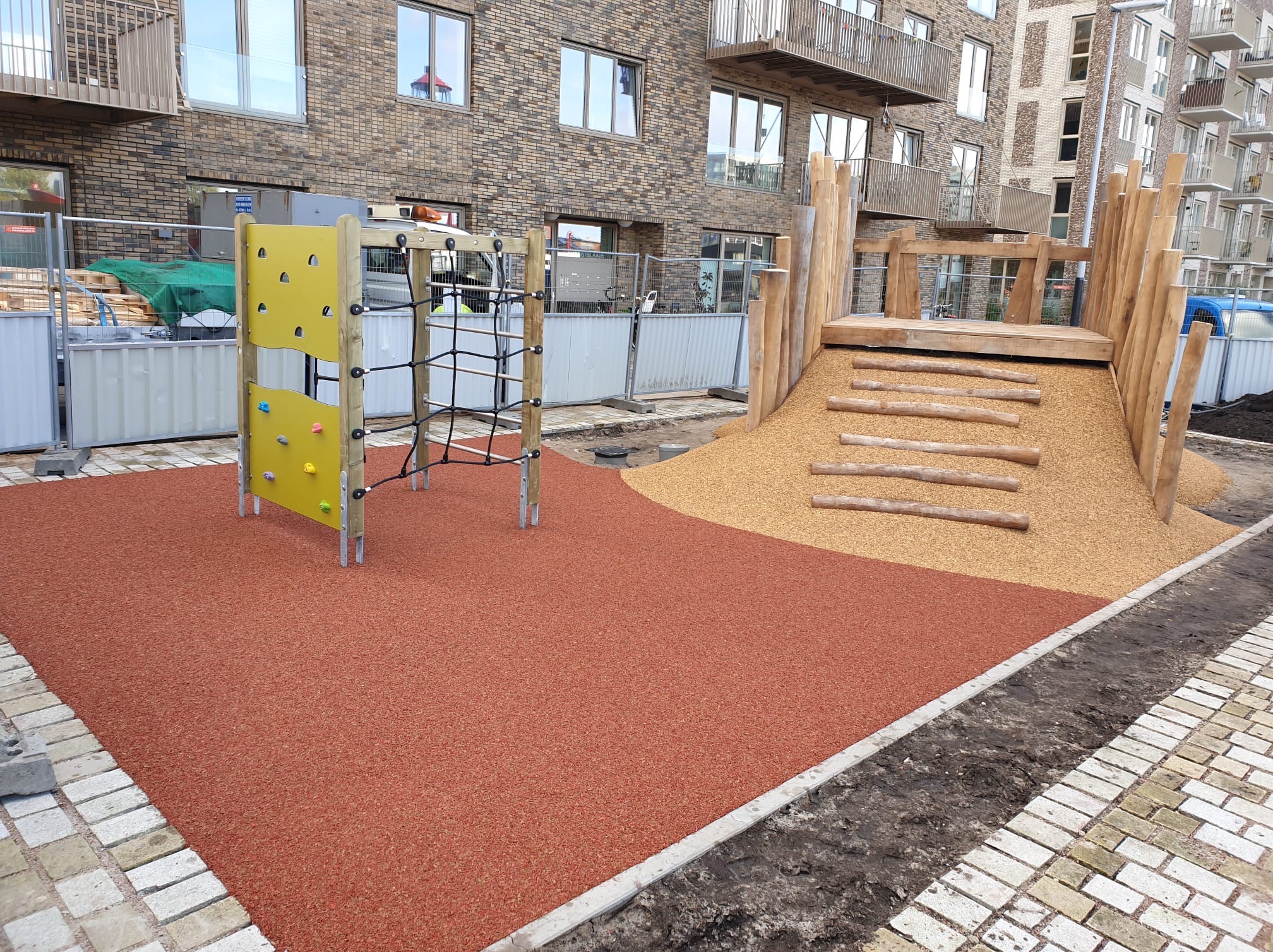 Nowoczesny plac zabaw z żółtą ścianką wspinaczkową i drewnianą konstrukcją ze zjeżdżalnią, wykończony czerwoną nawierzchnią bezpieczną.