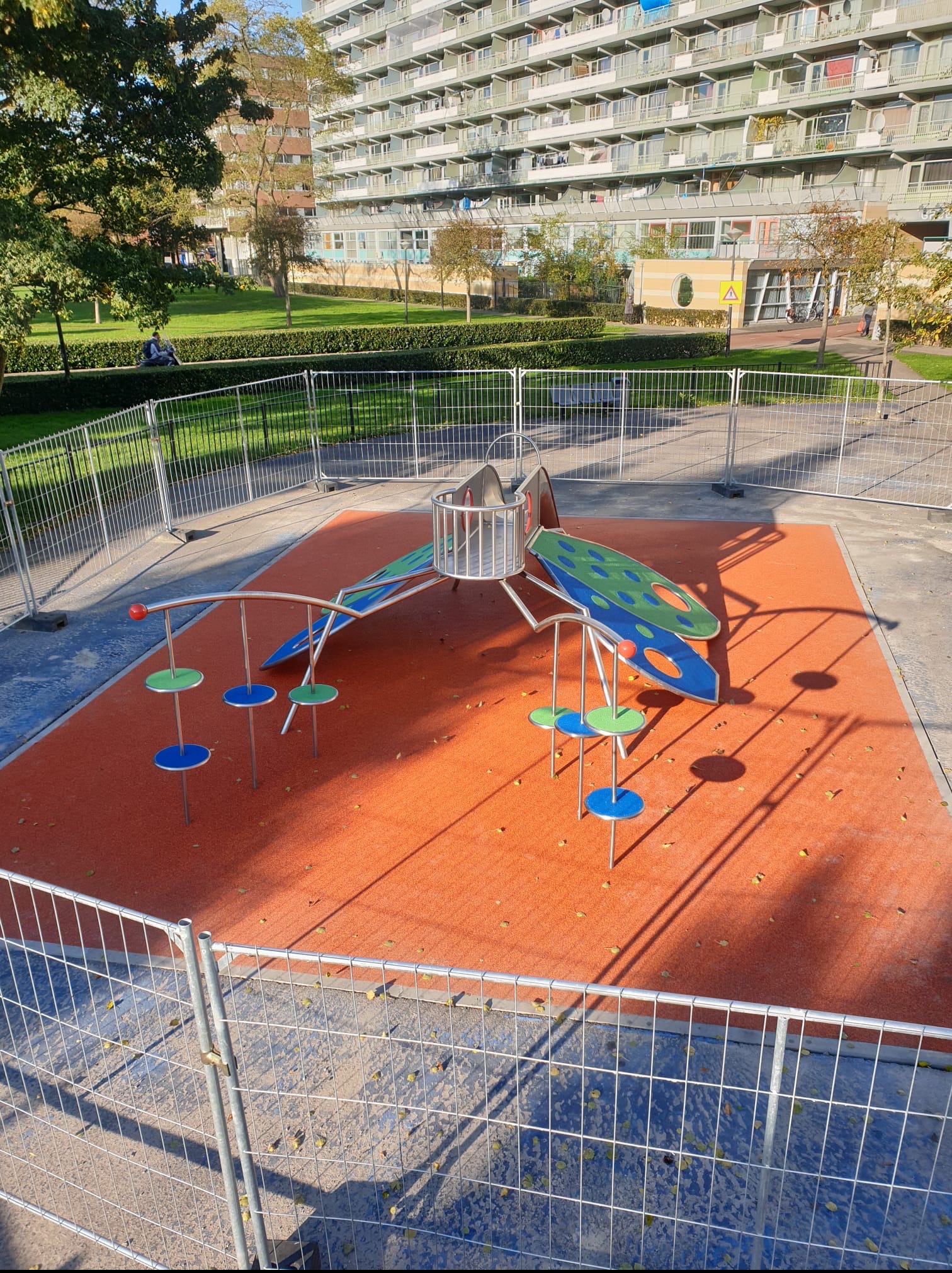 Nowoczesny plac zabaw z metalowymi elementami i pomarańczową nawierzchnią, otoczony tymczasowym ogrodzeniem, na tle wysokiego bloku mieszkalnego.