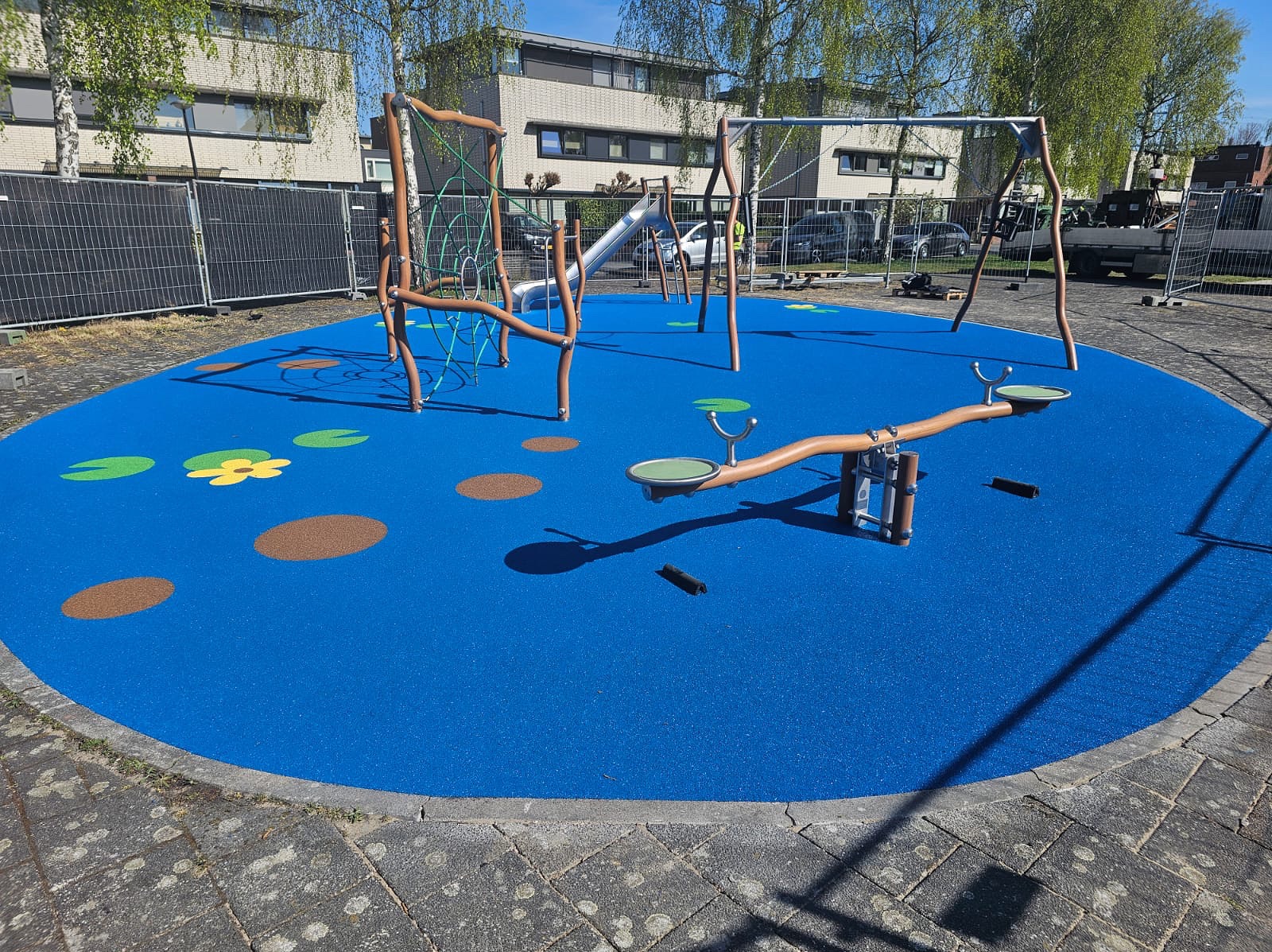 Niebieska nawierzchnia placu zabaw z huśtawką, zjeżdżalnią i drabinką linową. Na podłożu dekoracyjne brązowe i zielone elementy. W tle budynki mieszkalne.