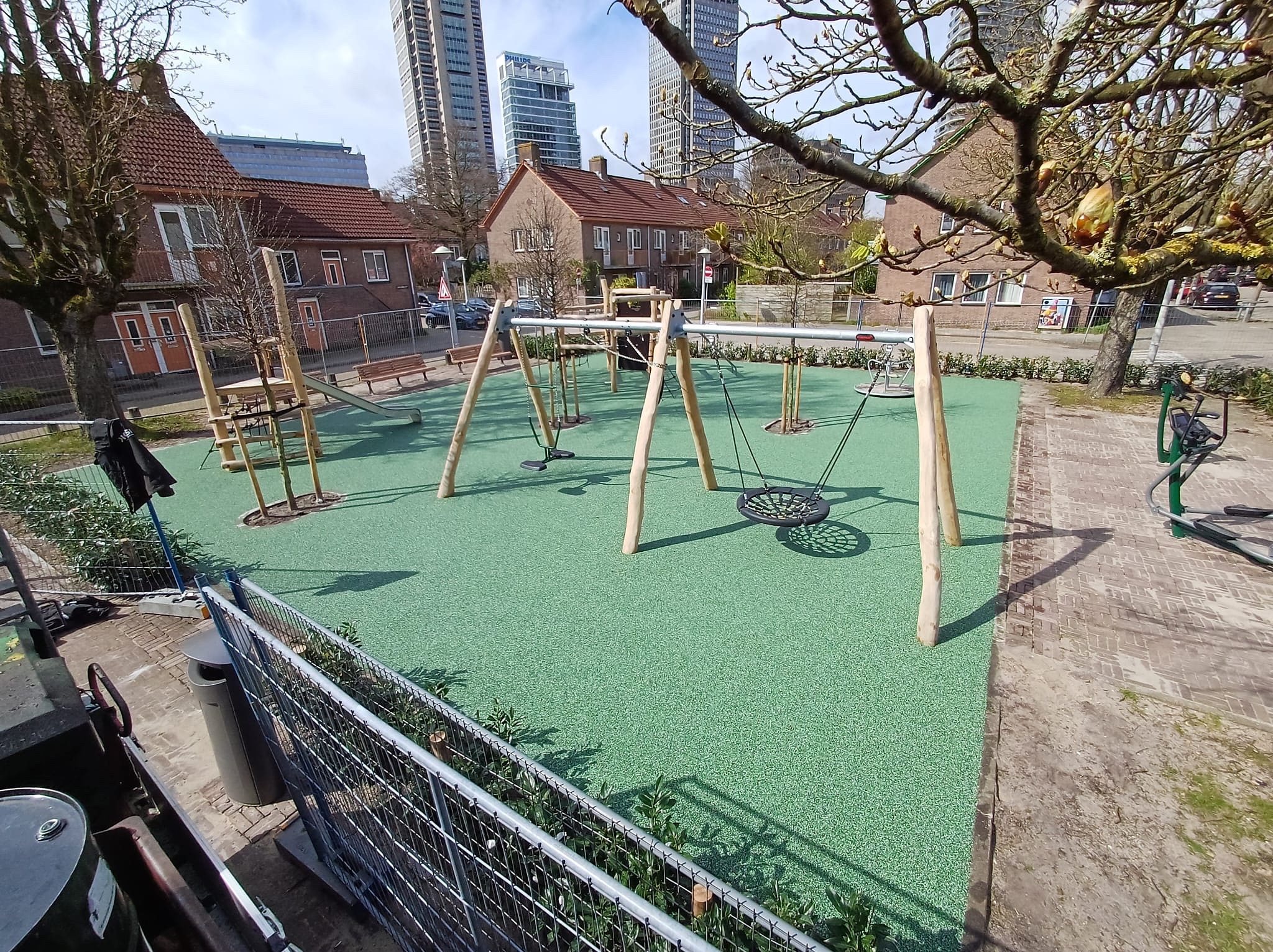 Nowoczesny plac zabaw z huśtawkami i drabinkami na zielonej nawierzchni, w tle budynki mieszkalne i wieżowce. Całość ujęta w słoneczny dzień.