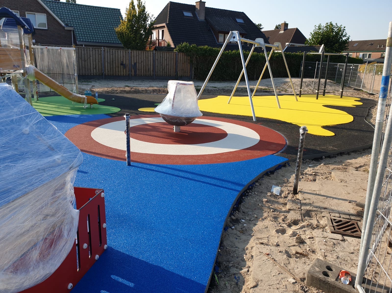 Nowo wybudowany, kolorowy plac zabaw z huśtawkami i karuzelą, jeszcze w folii ochronnej, otoczony piaskiem i ogrodzeniem tymczasowym.