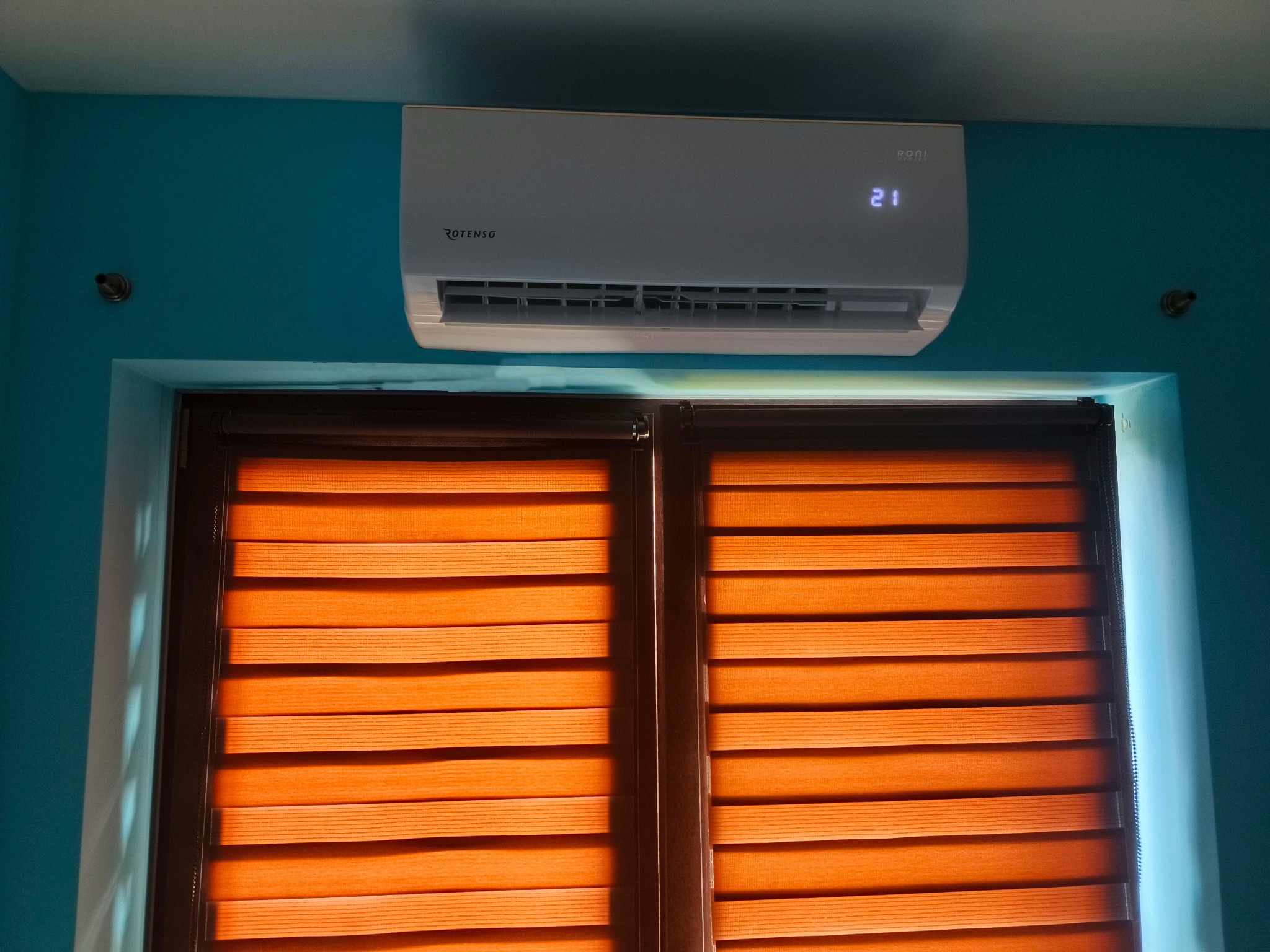Biała klimatyzacja Rotenso RONI 2.6 kW zamontowana na turkusowej ścianie nad oknem z pomarańczowymi roletami w poziomym ujęciu.