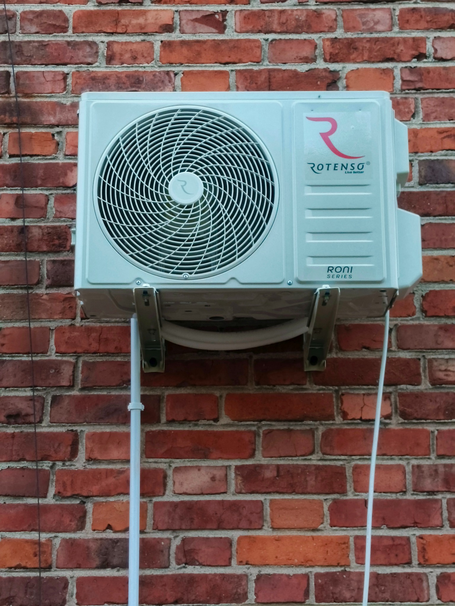 Zamontowana jednostka zewnętrzna klimatyzacji Rotenso RONI na czerwonej, ceglanej ścianie. Widoczne elementy montażowe i rurka odprowadzająca.