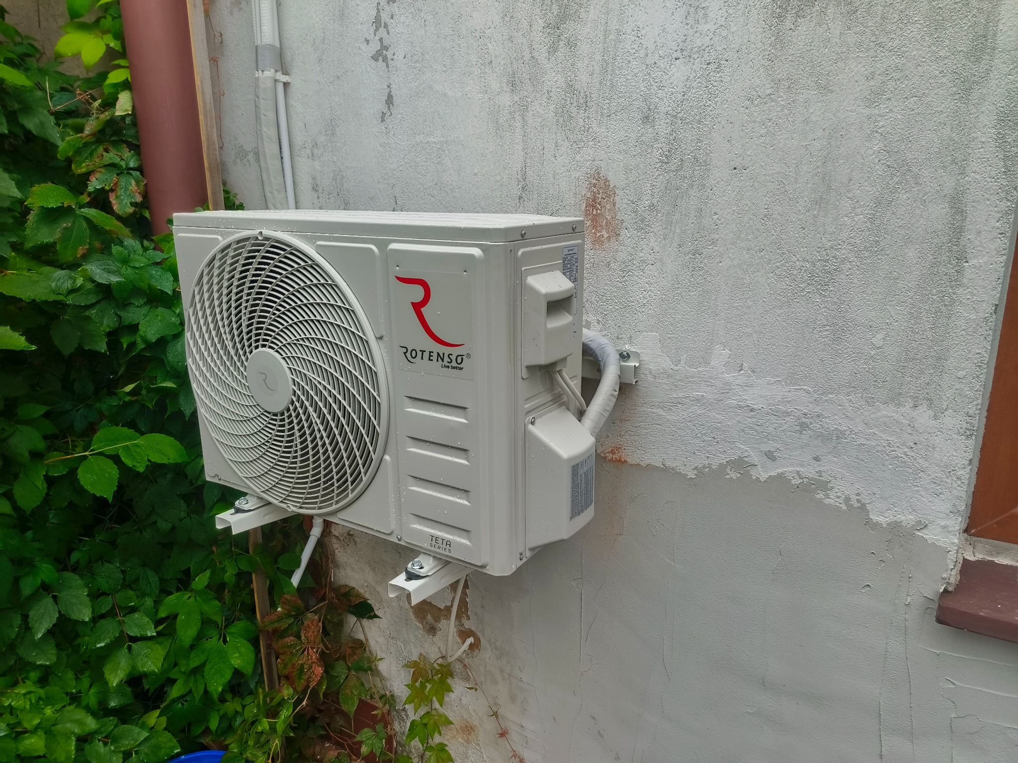 Biała jednostka zewnętrzna klimatyzacji Rotenso zamontowana na szarej, teksturowanej ścianie budynku z zielonymi pnączami u dołu, widoczne elementy instalacji.